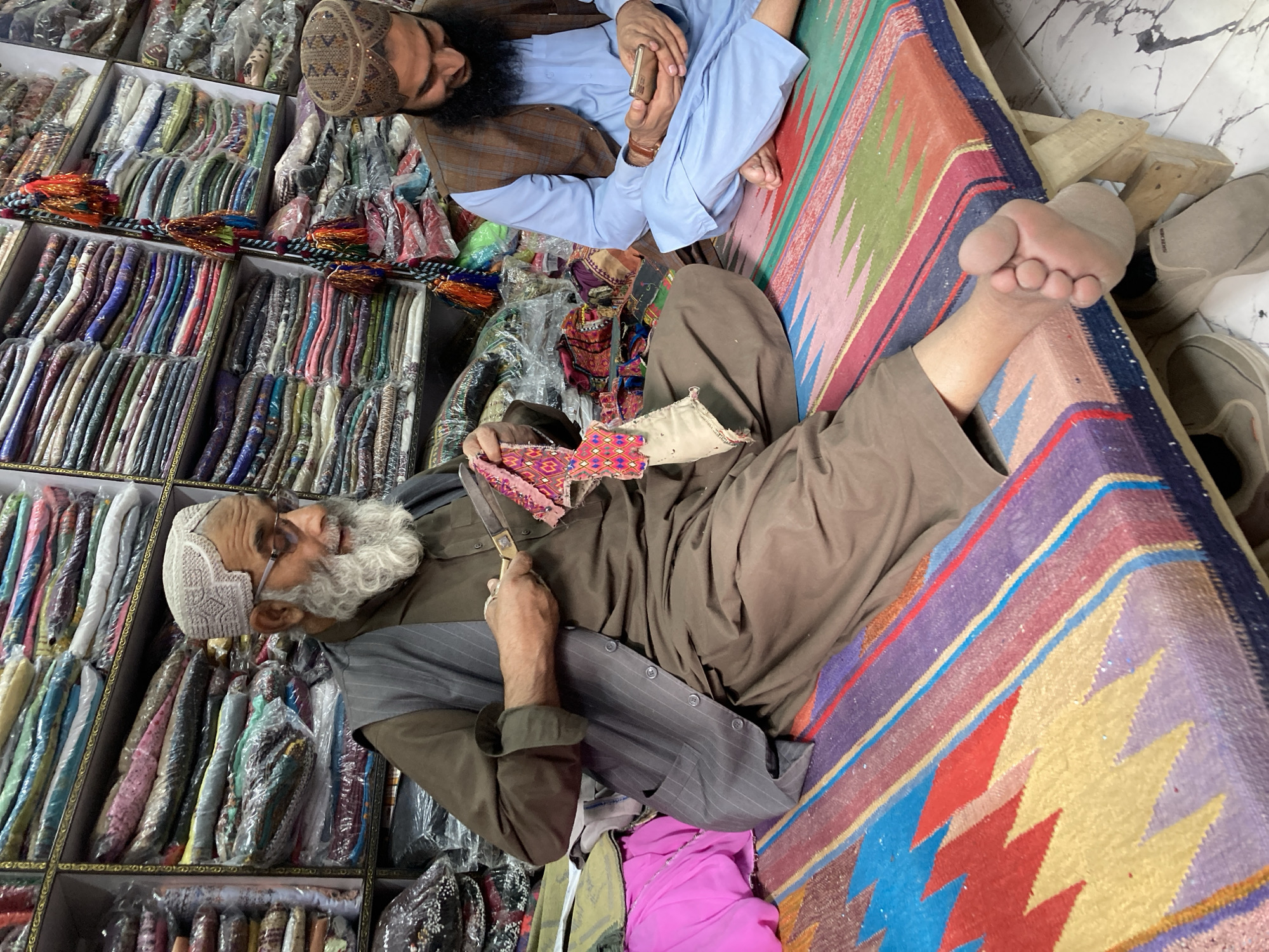 Two men are sitting on a colorful rug in what appears to be a shop in Quetta City Tehsil, Pakistan. One man, with a long white beard and wearing glasses and a patterned white cap, is seated with his legs extended. He holds a piece of patterned fabric in his left hand and a pair of scissors in his right hand. He is wearing a brown shirt with a grey pinstripe vest. The second man, with a dark beard and wearing a patterned brown cap, is seated cross-legged and is dressed in a light blue shirt and a brown vest. Shelves filled with fabrics and packaged goods are visible in the background. A bare foot and a pair of shoes are near the edge of the rug.
