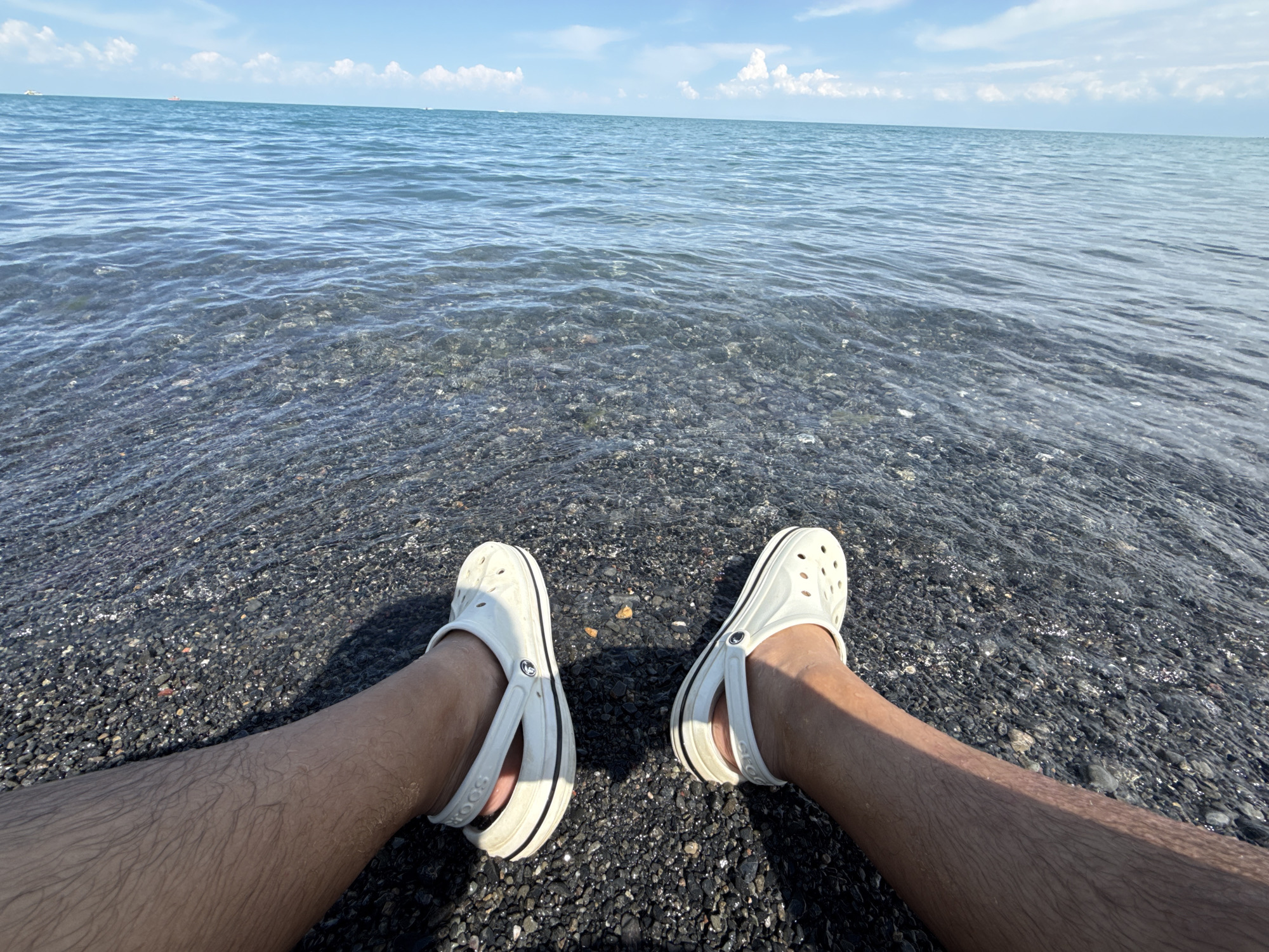 A first-person perspective shot shows a person's legs and feet, clad in white Crocs, resting on a dark, pebble-strewn beach. The water of a large body of water laps at the shore, creating ripples and reflecting the bright sky. The water is clear, allowing the pebbles beneath to be visible.
In the distance, the horizon is defined by the meeting of the water and the sky, dotted with a few small white clouds. The sky is a bright blue, suggesting a clear and sunny day. The overall impression is one of relaxation and tranquility, with the gentle sound of waves and the warmth of the sun implied. The presence of the beach and the large body of water suggests a coastal or lakeside setting. The location context provided, Ақши, Kazakhstan, indicates this is likely a scene from that region.
There is no visible text in the image. The time of day appears to be daytime, likely late morning or afternoon, given the bright sunlight. The weather is fair and sunny.