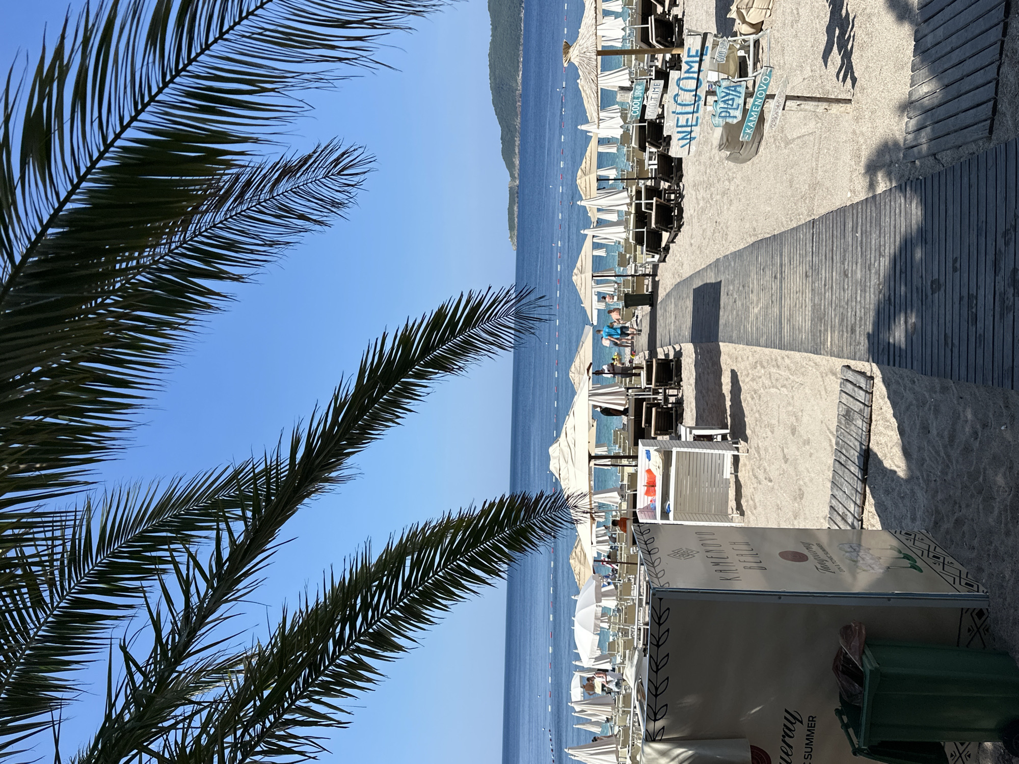 This image depicts a scene at Kamenovo Beach in Boreti, Montenegro. Palm fronds frame a view of a sandy beach with rows of empty lounge chairs and umbrellas. A wooden boardwalk leads towards the calm blue sea, where buoys mark a swimming area. In the foreground, a large advertising panel for "KAMENOVO BEACH" and "Tanqueray Gin & Tonic Summer" stands next to a green trash bin. Further along the beach, a signpost displays "WELCOME," "PLAYA," and other directional information. The sky is clear and blue.