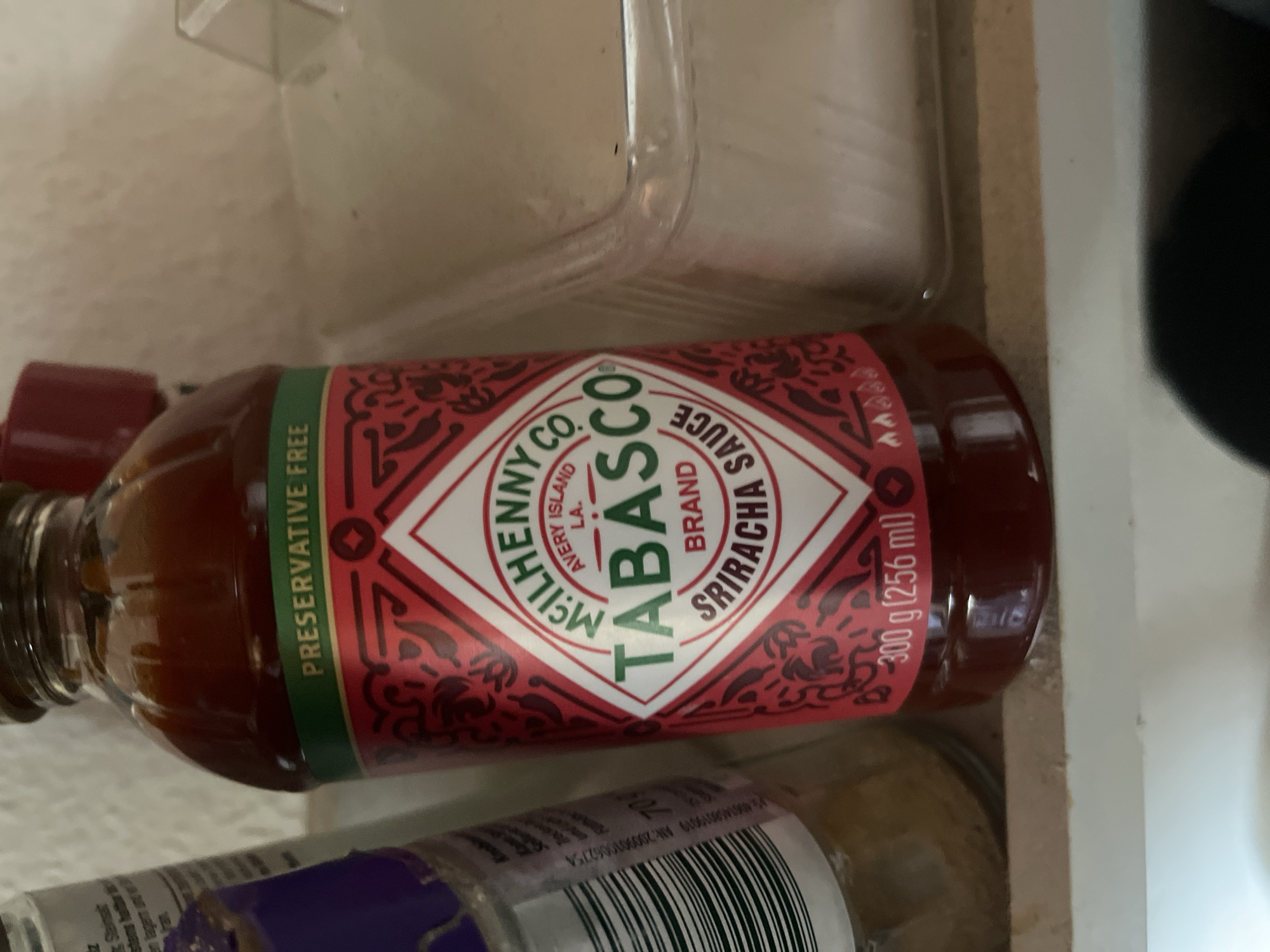 The image shows a bottle of Tabasco Sriracha sauce. The bottle is red with a green band that says "PRESERVATIVE FREE". The label has a diamond shape with "TABASCO" in large letters. There is text below it that says "BRAND SRIRACHA SAUCE". There is also text at the bottom of the bottle that says "300 g (256 ml)". To the left of the Tabasco bottle is another bottle with a purple cap and a white label with black text and a barcode. To the right of the Tabasco bottle is a clear plastic container. The items are on a shelf. There is no indication of the city of Schwäbisch Hall, Germany in the image.