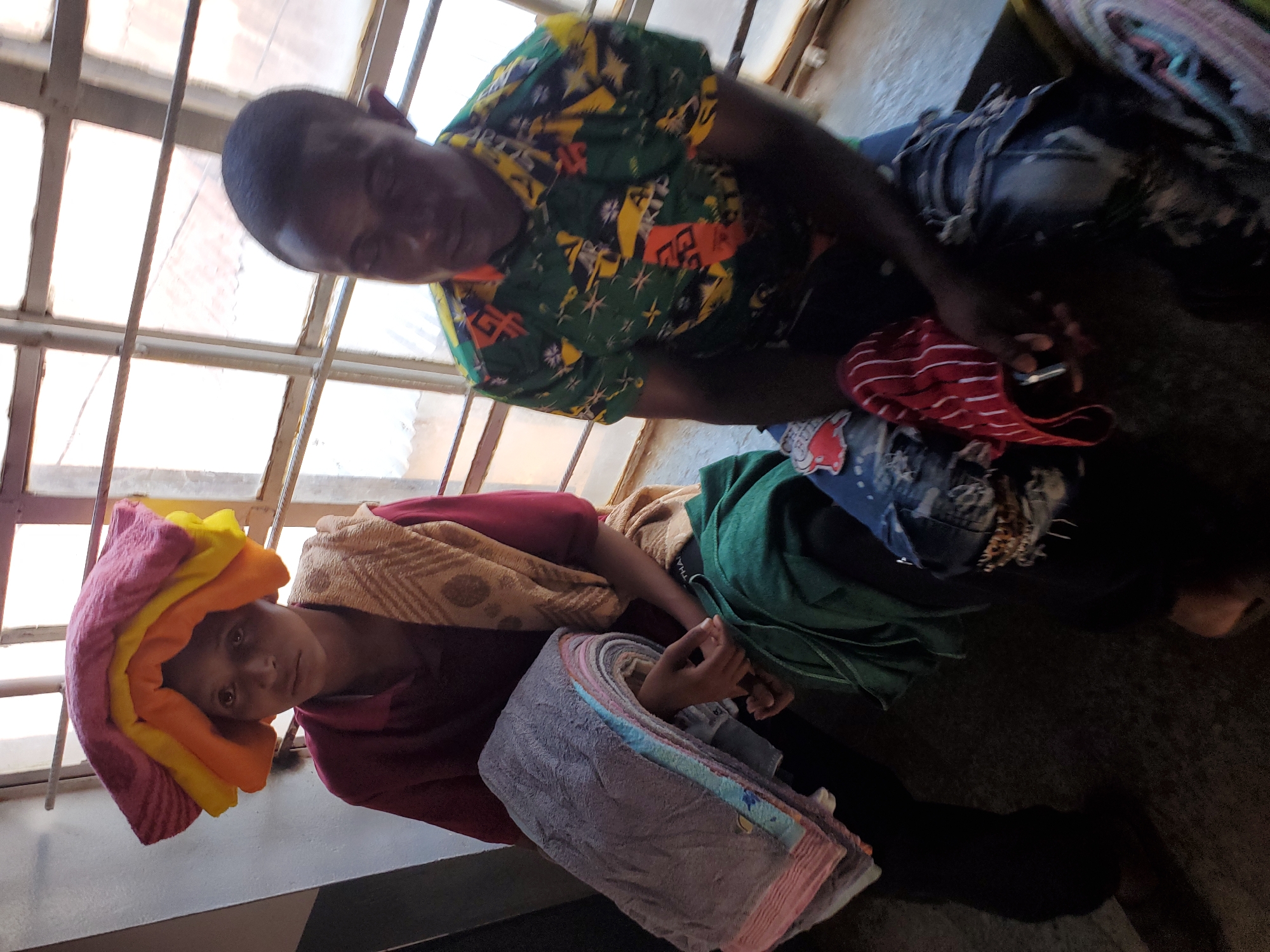 This indoor image, captured in Jos, Nigeria, features two individuals illuminated by natural light streaming through a barred window. The scene appears to depict an everyday activity related to textiles or laundry.
On the left, a young woman or girl looks directly at the viewer with a neutral expression. She wears a reddish-brown garment. Stacked on her head are multiple neatly folded towels in vibrant hues of pink, yellow, and orange. Her hands are positioned near a neat pile of pastel-colored folded cloths or towels, including shades of grey, blue, and light pink.
To the right, an adult man is seated, his gaze directed downwards towards an object in his lap. He wears a short-sleeved, brightly patterned shirt featuring green, yellow, orange, and black designs. His hand appears to be holding a red and white striped fabric item, possibly a garment, resting near denim jeans. In the background behind him, a colorful, possibly woven, mat or rug is partially visible.
The setting is an informal indoor space, likely a home or a small workshop, given the nature of the activity. A window with visible metal security bars spans the background, letting in ample daylight. The floor is dark and utilitarian. The overall impression is one of routine domestic or small-scale commercial activity involving the handling and organization of various textile items. No visible text is discernible in the image.
