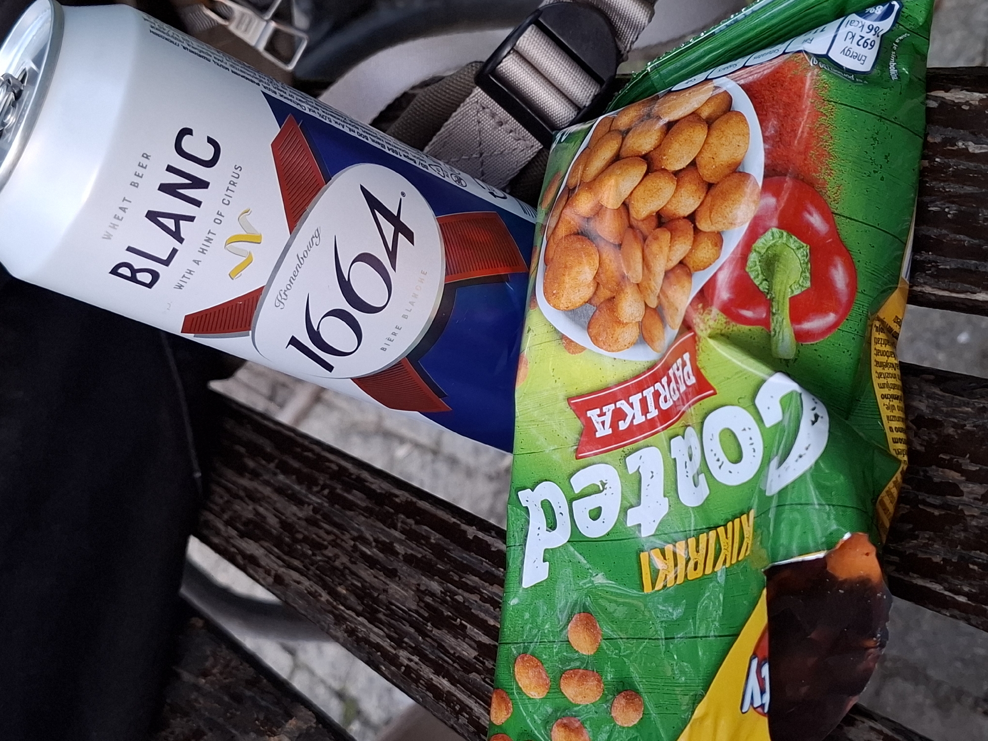 This outdoor still life image, captured in Novi Sad, Serbia, depicts a casual break on a wooden bench. The primary subjects are a can of Kronenbourg 1664 Blanc wheat beer and a bag of Kikiriki Coated Paprika peanuts.
The beer can, predominantly white with blue and red details, prominently features the text "WHEAT BEER BLANC WITH A HINT OF CITRUS" and the brand "Kronenbourg 1664 BIÈRE BLANCHE". Additional text includes "500 ml alc. 5.0% vol". The can's tab is visible, suggesting it's ready for consumption.
Next to the beer is a colorful green and yellow snack bag. It displays an image of numerous coated peanuts and a large red bell pepper, indicating its "PAPRIKA" flavor. Large white lettering spells "Coated KIKIRIKI" (Kikiriki is Serbian for peanut). Visible nutritional information states "Energy 692 kJ / 166 kcal". Other smaller text in Serbian/Croatian includes "ulje" (oil), "kikiriki", "inozitat", "karbonat", and "adržati" (to contain). Part of a "Party" logo is also visible at the bottom right.
These items are placed on a dark brown, weathered wooden bench, characteristic of outdoor public seating. A blurred grey pavement is visible in the background, suggesting a park or urban outdoor area. A beige/grey backpack strap with a black plastic buckle is partially visible behind the items, implying they were recently carried. The natural lighting indicates it is daytime. The scene conveys a relaxed moment, enjoying snacks and a drink outdoors.
