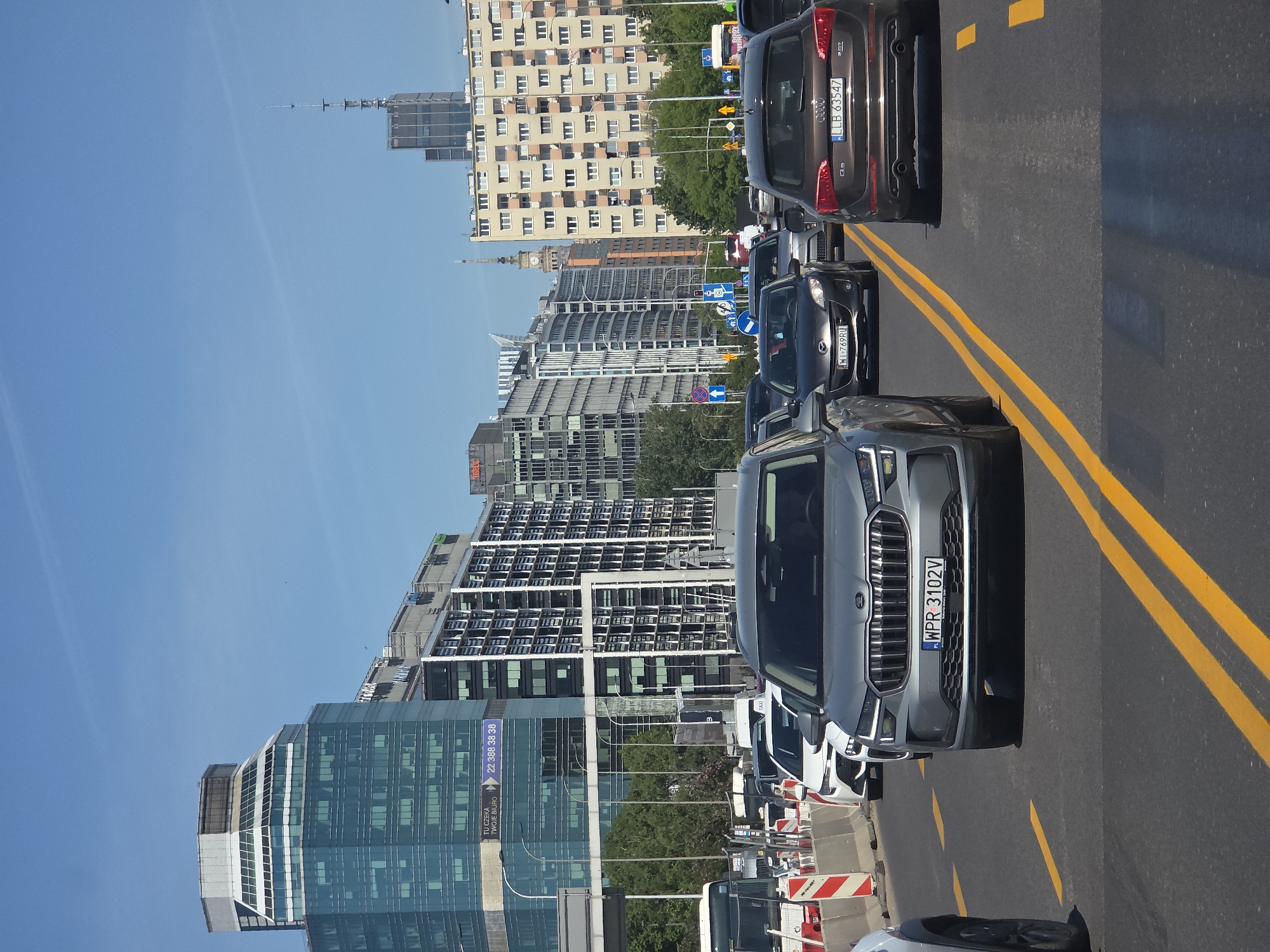The image shows a street scene in Warsaw, Poland, with a line of cars stopped in traffic. The buildings in the background are a mix of modern skyscrapers and older, more traditional buildings, including the Palace of Culture and Science. The sky is clear and blue. The road has double yellow lines indicating no passing, and construction barriers are present near the left edge of the image. The license plate WPR 3102V is visible on the front of a gray car. Another car displays the license plate LLB 63547. The number 22 388 38 38 is visible on a building banner, advertising office space.