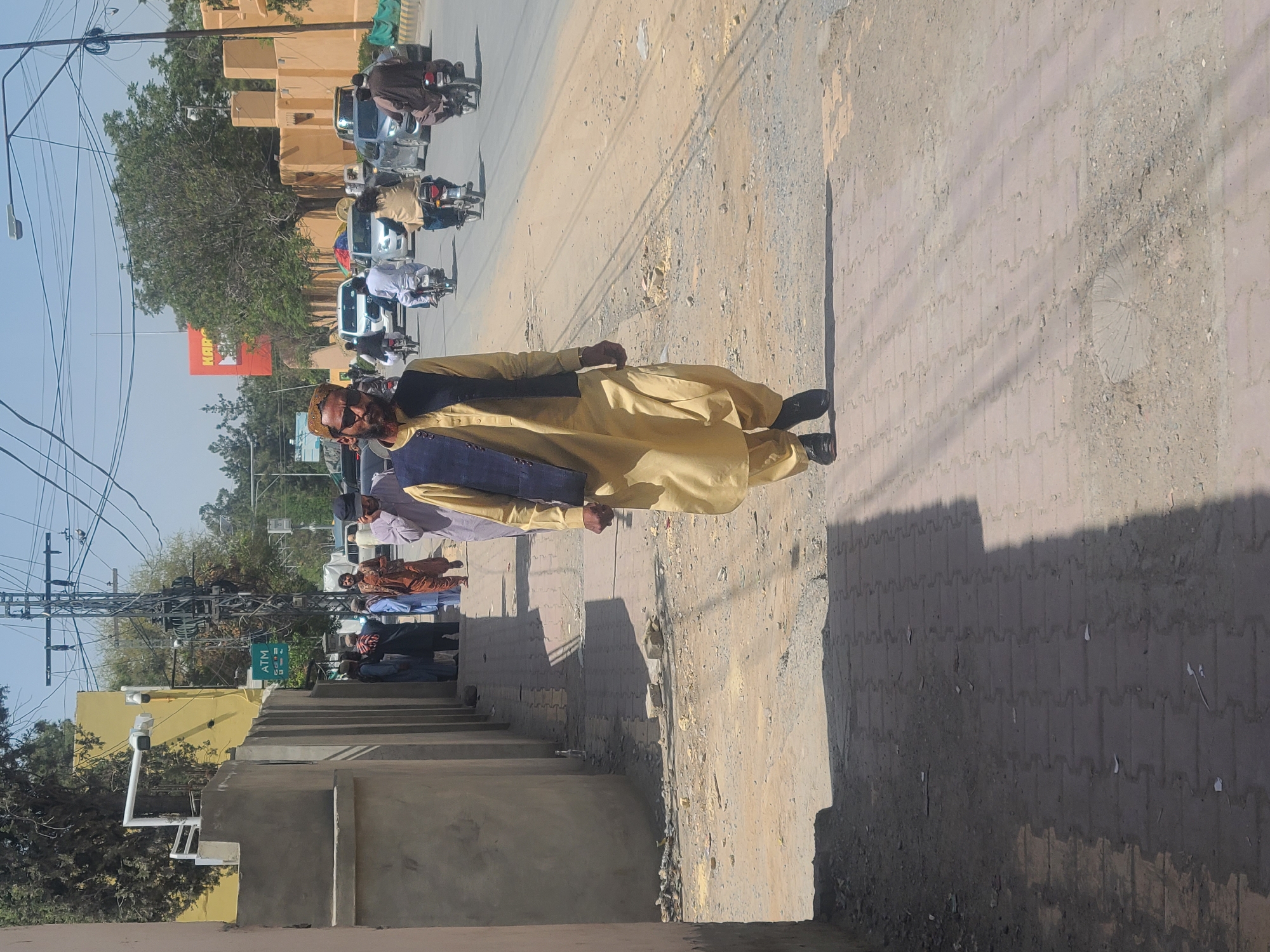 A sidewalk in Quetta City Tehsil, Pakistan features a man in a yellow shalwar kameez and dark vest standing on the sidewalk next to a street with vehicle traffic. The man has a beard and dark sunglasses. Other people stand in the background near a building. Overhead power lines crisscross above the street. A sign with the word "KAR" is visible in the background.