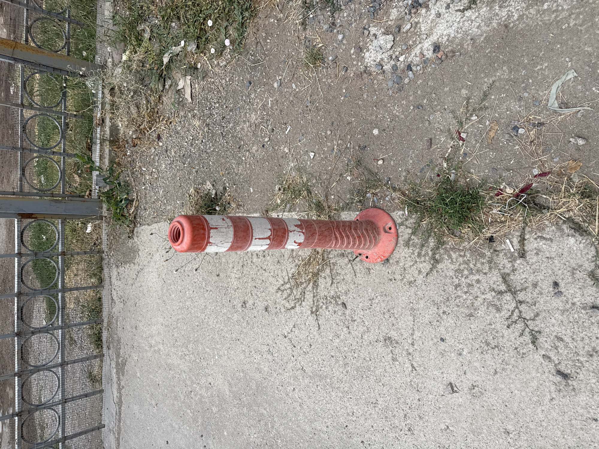 The image shows a red and white traffic cone on a patch of dirt and gravel next to a concrete sidewalk. A metal fence with circular designs is visible in the background. There is sparse vegetation and some litter on the ground. The scene appears to be an outdoor urban environment in Yangibazar, Uzbekistan.