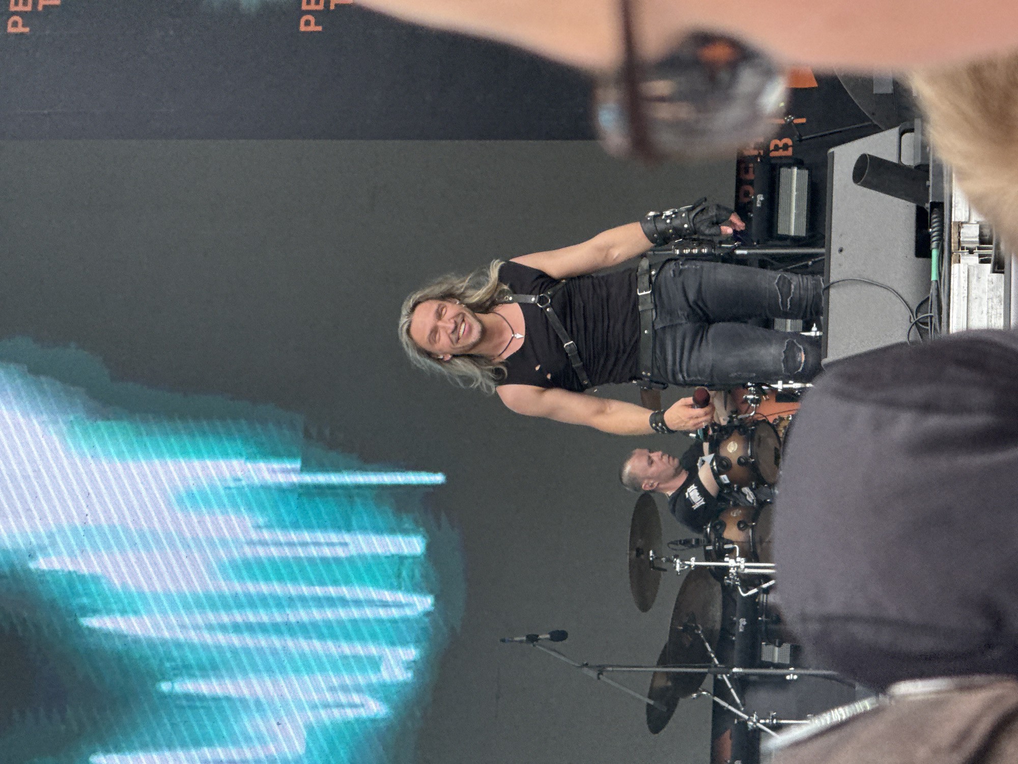 A male performer with long blonde hair and a black sleeveless shirt with harness details is smiling and looking at the camera on a stage. He is wearing ripped black jeans and has a microphone stand and a drum set behind him. The background features a large screen displaying a light blue, abstract pattern. The scene appears to be an outdoor music event. There are spectators in the foreground, partially obscuring the view. Text is visible on a black backdrop to the right, with the letters "PE" and "T" appearing to be part of a larger word. This event is taking place in Lagunovo, Russia.
