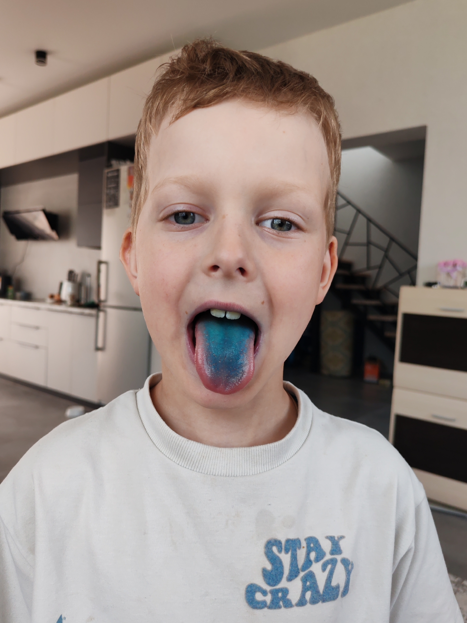 A young Caucasian boy with light reddish-blond hair and blue eyes is shown from the chest up, facing forward, with his mouth open to reveal a blue-stained tongue. He wears a white crew neck t-shirt with the words "STAY CRAZY" in blue on the front. He is in what appears to be a modern kitchen or dining area with white cabinets and a staircase visible in the background. The location is not explicitly provided in the image and while the question asks for the location to be in Chaikovsky, Russia, the image itself does not provide enough information to pinpoint the location accurately.