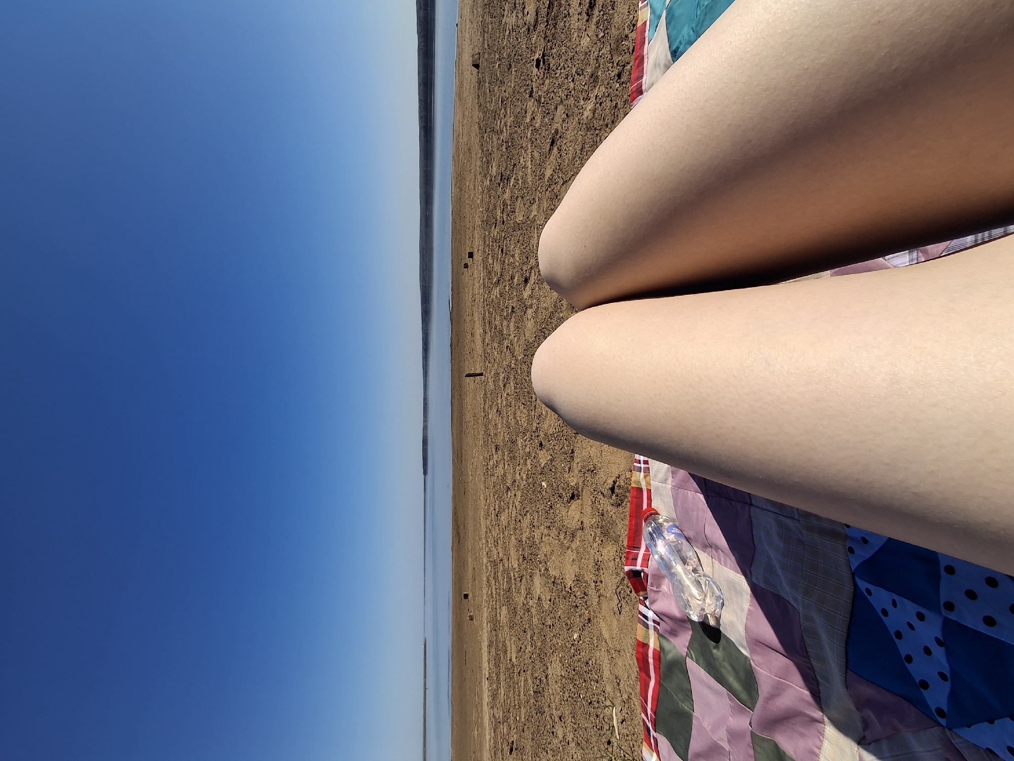 A first-person perspective shot shows a pair of legs resting on a patchwork quilt, suggesting someone is lounging on a beach. The beach is sandy with some debris scattered around. To the left of the frame is a view of a body of water, presumably a large river or reservoir, with a strip of land visible on the opposite shore. The sky above the water is a clear, vibrant blue. There is a plastic water bottle near the legs. The location is likely in the city of Chaikovsky, Russia, given the prompt.