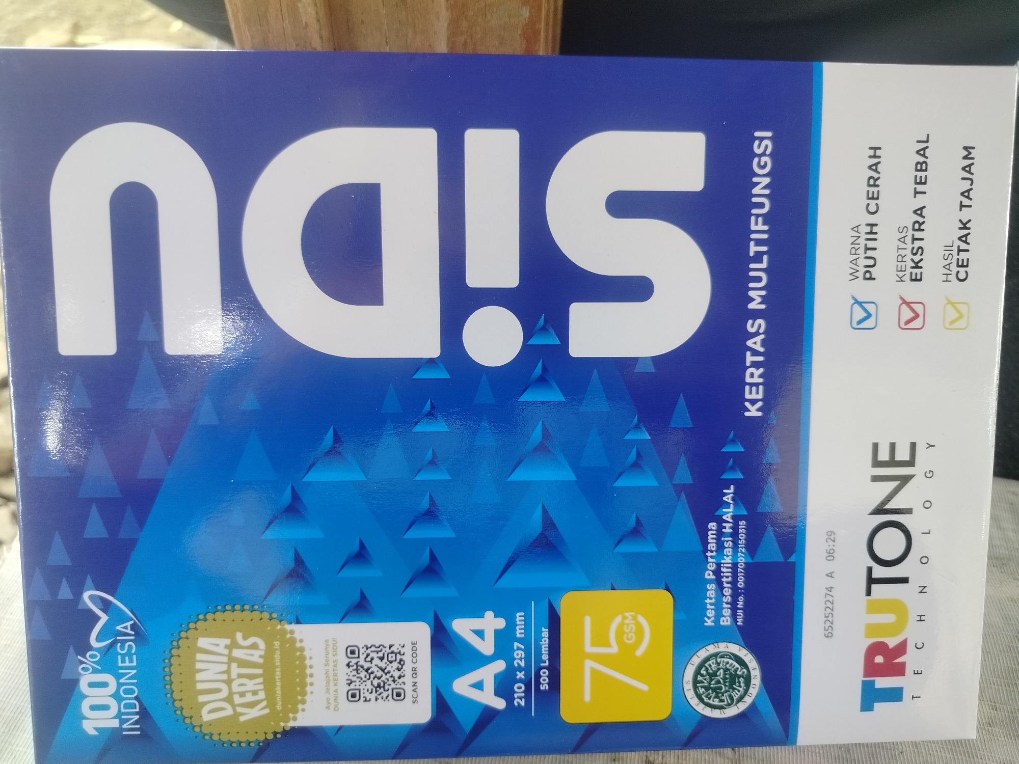 The image shows a close-up of a blue and white box of SIDU paper. The SIDU logo, large white letters forming "n.d.u.s!", is prominently displayed across the top of the box. To the right of the logo, there is text indicating "KERTAS MULTIFUNGSI" (Multipurpose Paper). Below this, there are checkboxes next to "WARNA PUTIH CERAH" (Bright White Color), "KERTAS EKSTRA TEBAL" (Extra Thick Paper), and "HASIL CETAK TAJAM" (Sharp Print Result).
On the left side of the box, there is a "100% INDONESIA" logo. Below this is a circular yellow graphic with the text "DUNIA KERTAS" (Paper World) and "duniakertas.sid.id". A QR code is also visible to the right of "DUNIA KERTAS".
The bottom of the box shows "A4 210 x 297 mm 500 Lembar" (500 Sheets), indicating the paper size and quantity. A yellow square with "75 GSM" is also present. Further to the right, there is a halal certification logo from MUI (Majelis Ulama Indonesia), and text stating "Kertas Pertama Bersertifikasi HALAL" (First Paper Halal Certified) with a MUI number.
The brand name "TRUTONE TECHNOLOGY" is displayed vertically on the right side of the box. The image is taken from an elevated, slightly angled perspective, suggesting it might be on a table or counter. The background is blurred, showing a wooden surface and hints of greenery, possibly outdoors.
There are no people or specific city locations identifiable in the image. The context of "Purwodadi, Indonesia" is not present within the visual information of the photograph.