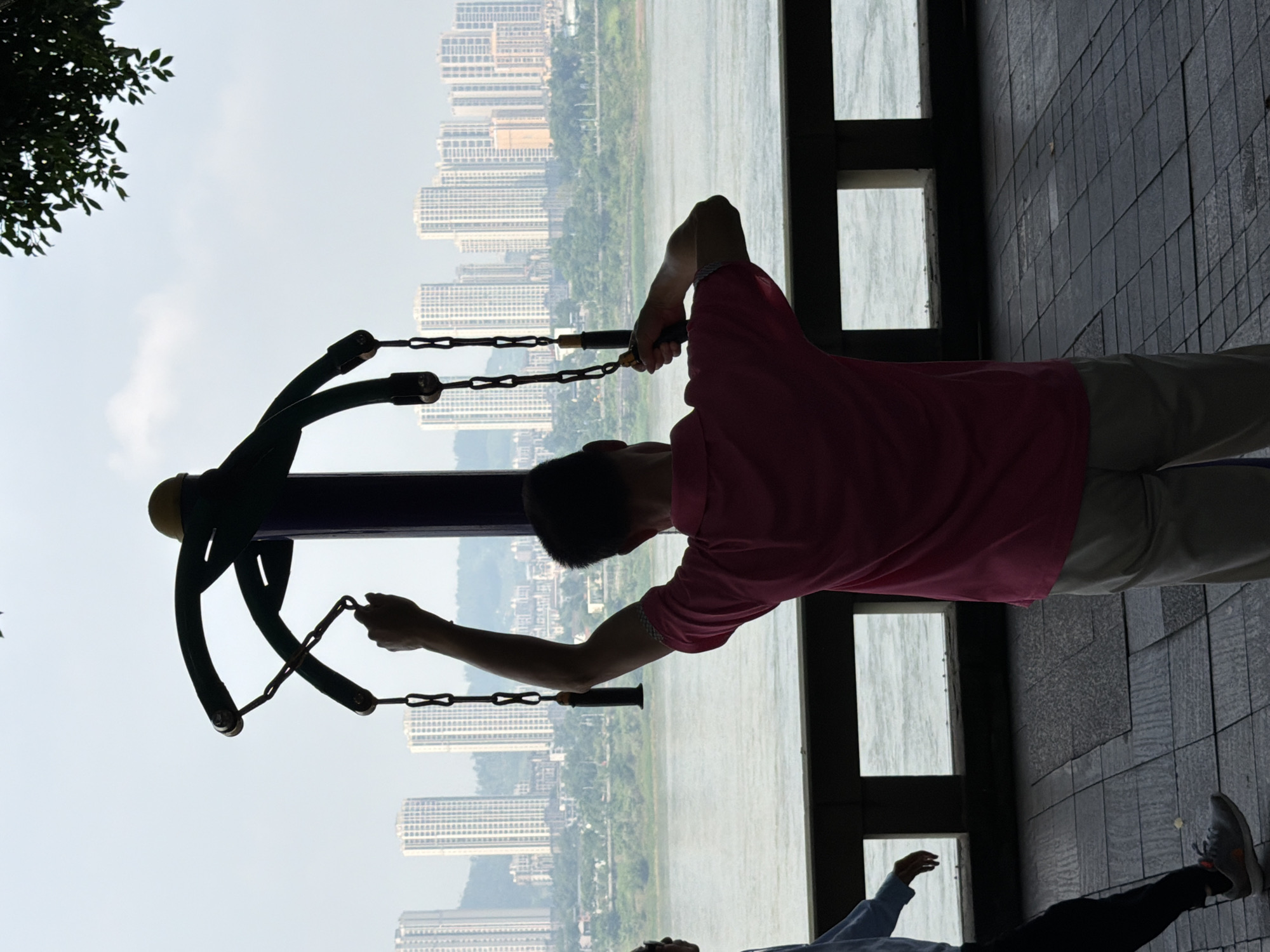This is a full, vertical shot of a man exercising on an outdoor gym machine, with a cityscape in the background. The man, silhouetted against the bright sky, is wearing a pink shirt and tan pants. He is holding onto the handles of the gym equipment, which is a large, dark green and purple structure with chains. His right arm is raised, and his left arm is extended forward. The gym equipment is positioned in front of a railing, beyond which is a wide body of water, likely the Jialing River. In the distance, a dense cityscape of tall, modern buildings is visible, with hills and trees interspersed. A portion of another person, wearing a blue shirt and black pants, is visible on the left side of the frame, walking away from the viewer. The foreground consists of a tiled walkway. The overall lighting suggests it is daytime, possibly late morning or early afternoon, with a slightly overcast sky. The image appears to be taken from the Jiangjin City Riverside Park, a popular recreational area in Jiangjin District, Chongqing, China, known for its river views and public exercise facilities.