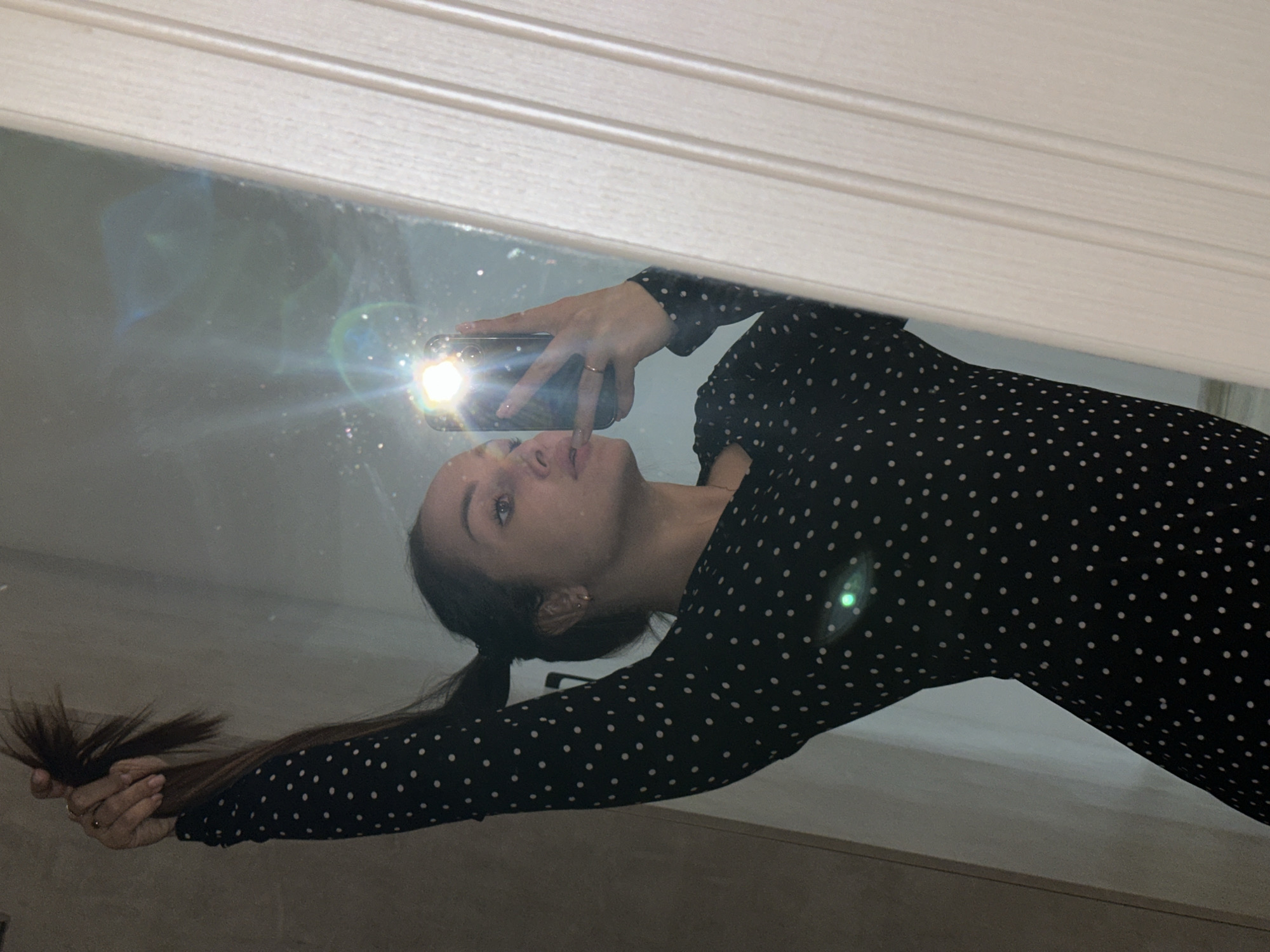 The horizontally oriented image, taken in Chaikovsky, Russia, captures a young woman's reflection in a mirror. She is holding a smartphone with her right hand, taking a selfie, and the device's bright flash intensely illuminates her face and the phone itself. Multiple prominent lens flares are visible on the mirror surface due to the flash.
The woman has long, dark hair, pulled back into a low ponytail or braid, with a section held in her left hand. She is wearing a long-sleeved, black dress covered in small white polka dots. A simple gold-colored ring is visible on her right ring finger, and her fingernails are painted a light, nude shade. She looks towards the phone with a neutral expression.
The setting appears to be an indoor space. Above the mirror's reflection, a light-colored, possibly wooden, paneled surface is visible. Below, a darker, indistinct surface, perhaps furniture or a counter, can be seen. The mirror itself appears to have some dust or speckles, particularly noticeable around the bright light sources. The ambient lighting of the room seems dim, suggesting the photo might have been taken at night or in a poorly lit environment. There is no visible text in the image.