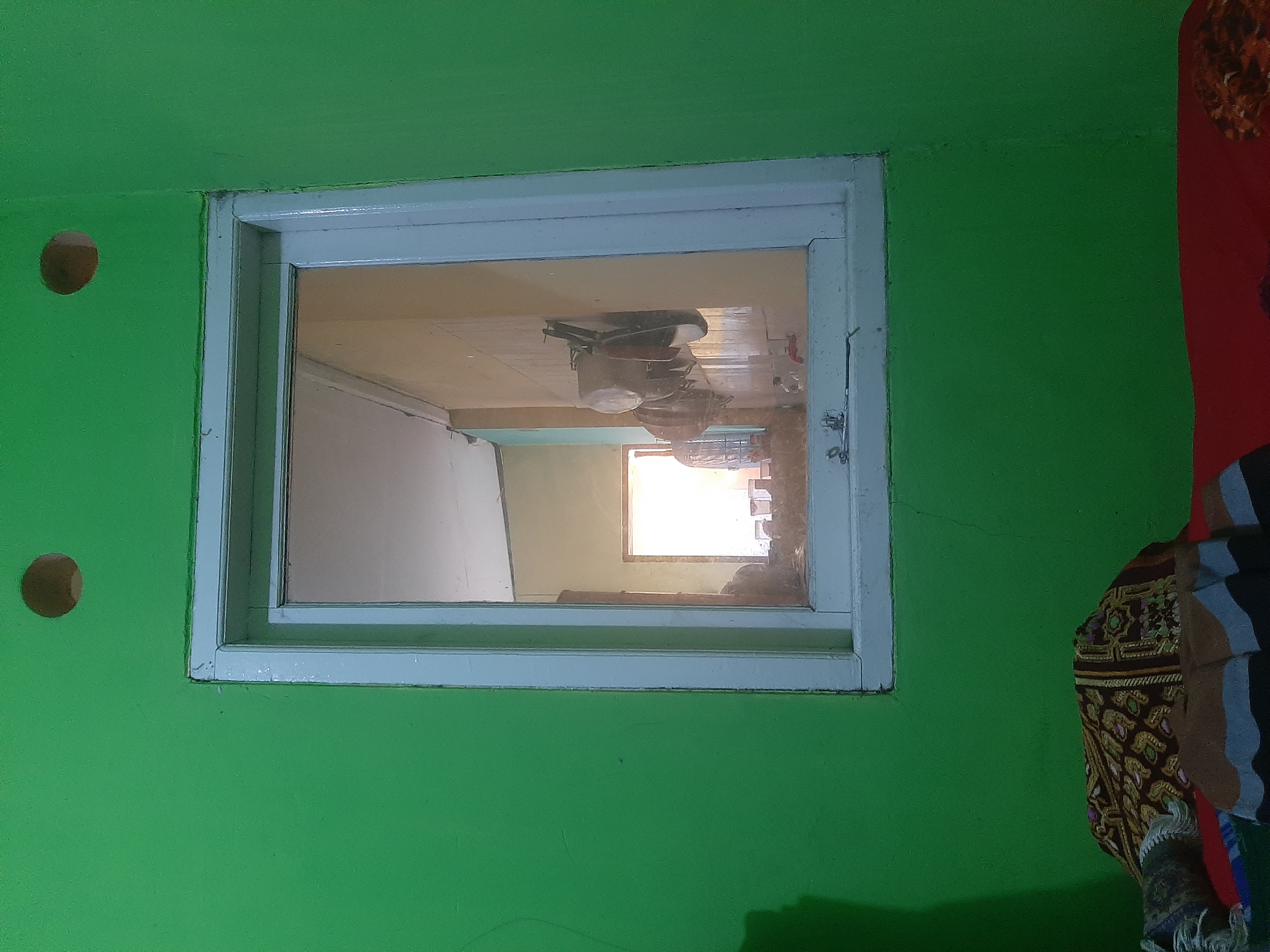 The image depicts an interior scene of a room, likely a bedroom, with a prominent green painted wall. A rectangular window with a white frame is centered on the wall. Through the window, another room is visible, including a doorway leading to a brightly lit space, and kitchen utensils hanging on a wall. Below the window, on the green wall, the corner of a bed is visible, covered with patterned blankets and fabric. Two circular holes are present in the green wall, one above the left side of the window and another above the right side. The image was taken at night in Sumampir, Indonesia.