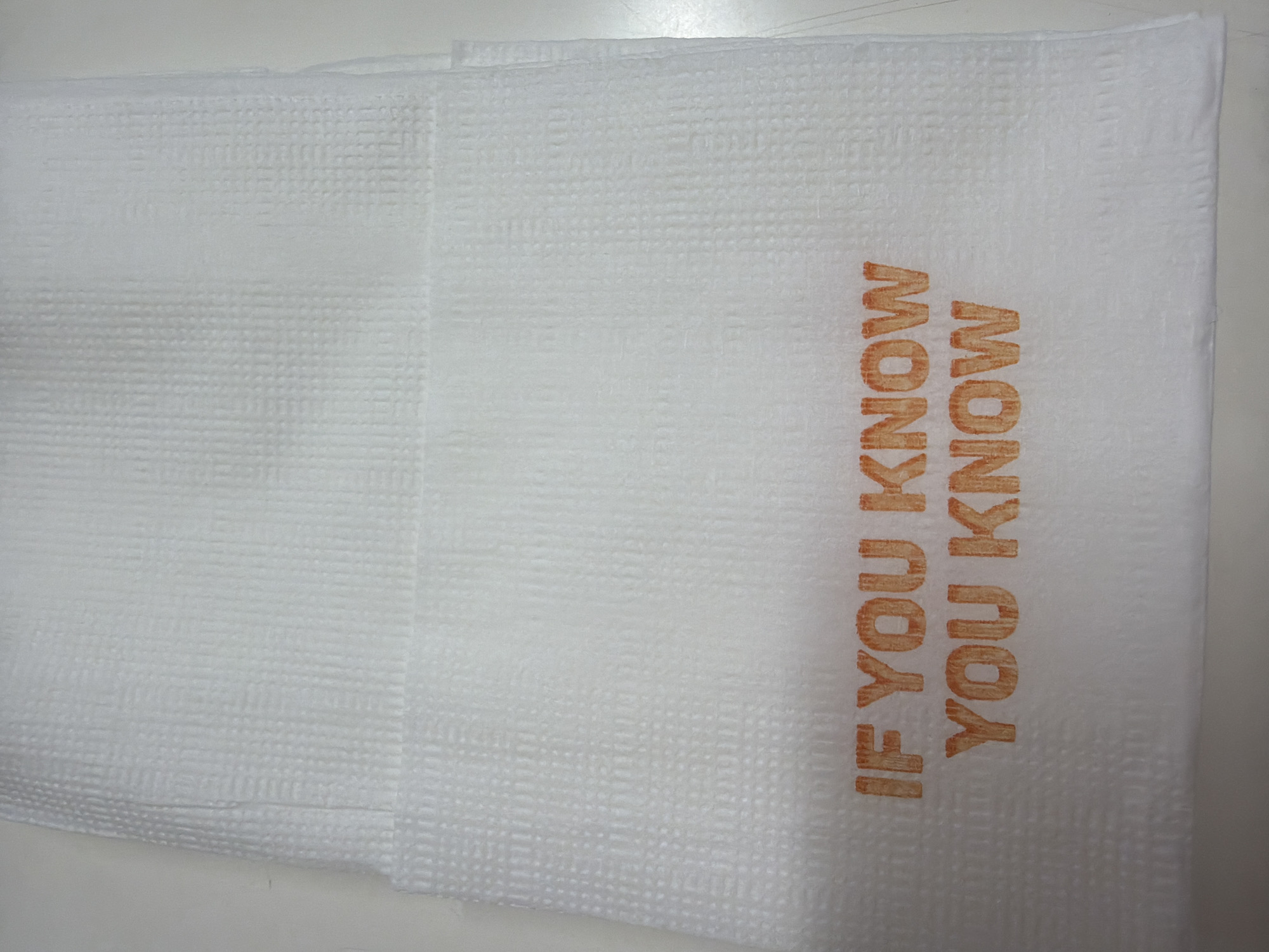 The image shows a stack of white paper napkins. The words "IF YOU KNOW YOU KNOW" are printed vertically in brown ink on the top napkin. The surface beneath the napkins appears to be a light-colored, possibly white, table. The location is unknown.