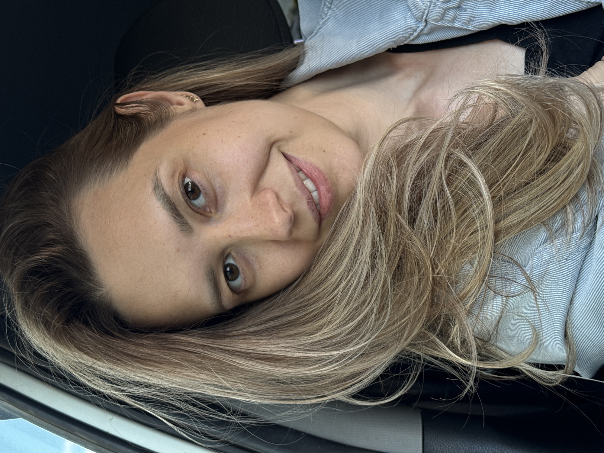 A casual portrait features a woman with long, wavy, light brown and blonde hair, which cascades around her face and shoulders. She has brown eyes, a fair complexion with a scattering of freckles, and light pink lips curved into a gentle, relaxed smile. Her right ear is adorned with small, star-shaped earrings.
She is dressed in a light blue, subtly striped or denim-textured top worn over a dark inner garment. The woman is positioned in what appears to be the interior of a vehicle, possibly a car, indicated by the dark upholstery of a seat or headrest behind her and the dark trim visible along the bottom edge of the frame. The lighting suggests it is daytime, with soft, indirect illumination on her face.
The activity taking place is a relaxed self-portrait or casual photograph, with the woman looking directly at the camera. Her expression conveys a sense of contentment and friendliness. The image is tilted, giving a dynamic angle to the composition. No visible text is present within the frame. The location context provided is Chaikovsky, Russia.