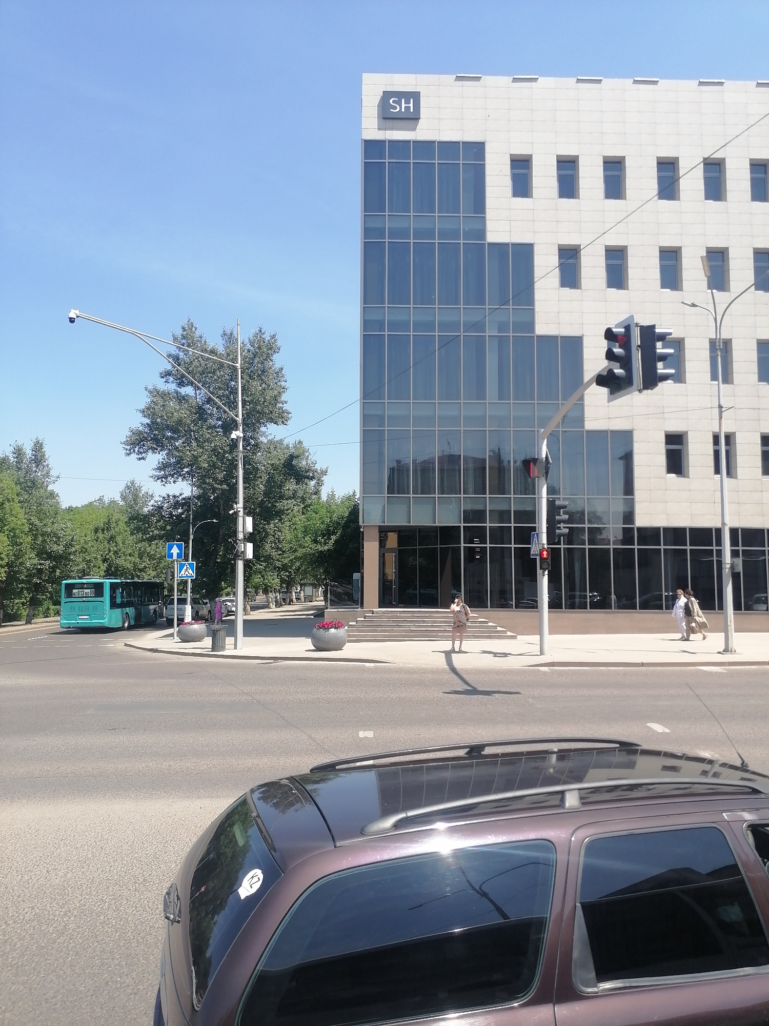 The image depicts an outdoor urban scene on a sunny day. In the foreground, the rear of a dark purple station wagon is visible. Beyond the car, a street runs horizontally across the frame. A blue public bus is traveling on the street from left to right. To the right of the street, a sidewalk is present with several people walking. A modern office building with a glass facade and light-colored stone accents stands prominently in the background. The building has a large "SH" logo at the top. Traffic lights are visible at an intersection. Trees line the street and sidewalks, adding greenery to the urban environment. The sky is clear and blue.