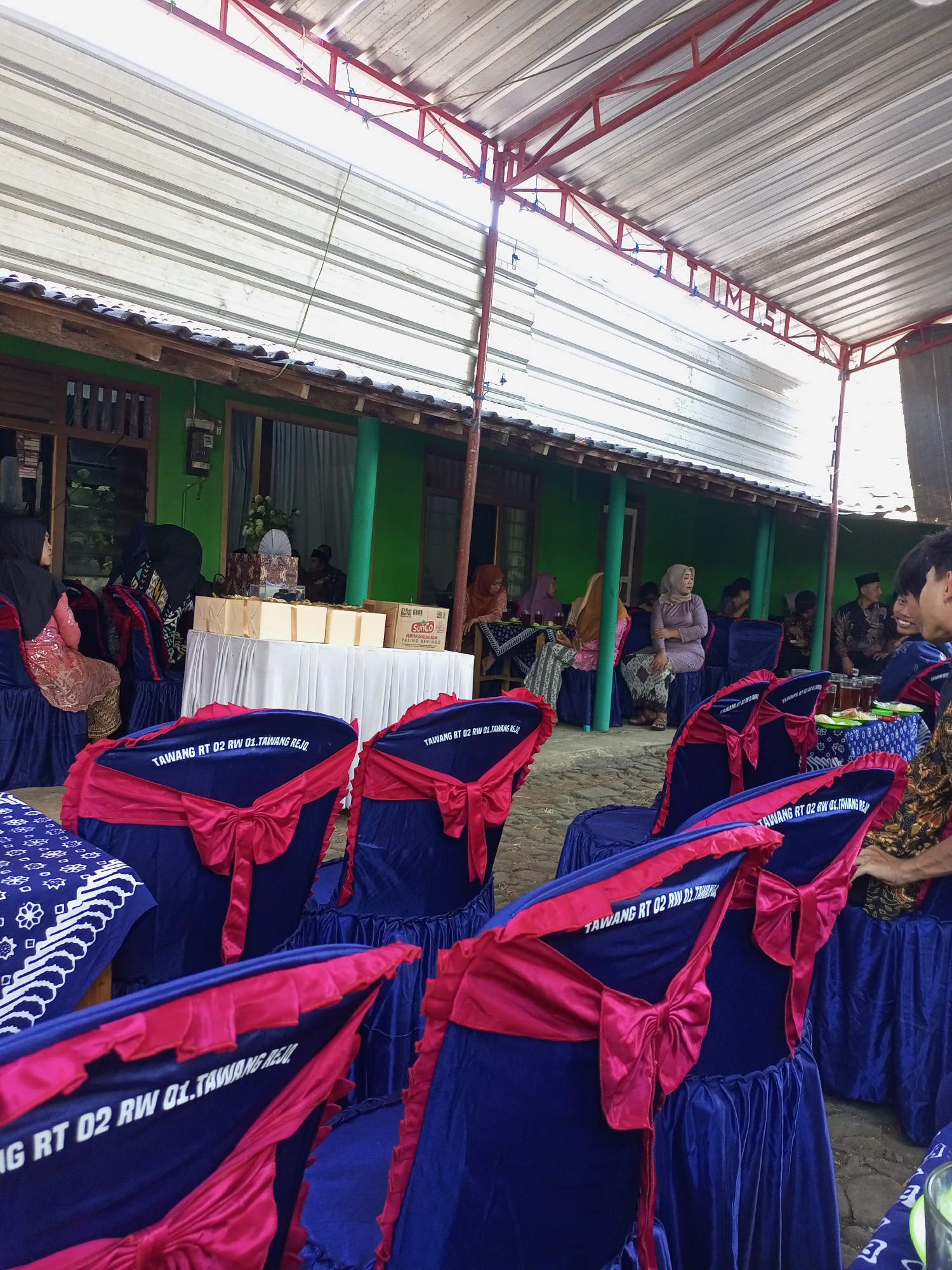 In Jatisrono, Indonesia, an outdoor event is taking place under a covered patio. The scene is dominated by rows of blue chairs adorned with bright pink sashes tied into bows. Several chairs in the foreground display white text that reads "TAWANG RT 02 RW 01.TAWANG REJO.". In the background, a green building with wooden window frames is visible. A table covered with a white cloth is set up, holding several boxes and what appears to be a decorative fan. A few individuals are seated at tables in the background, and others are standing, some engaged in conversation. The ground is paved with rough stones. The overall impression is of a community gathering or celebration.