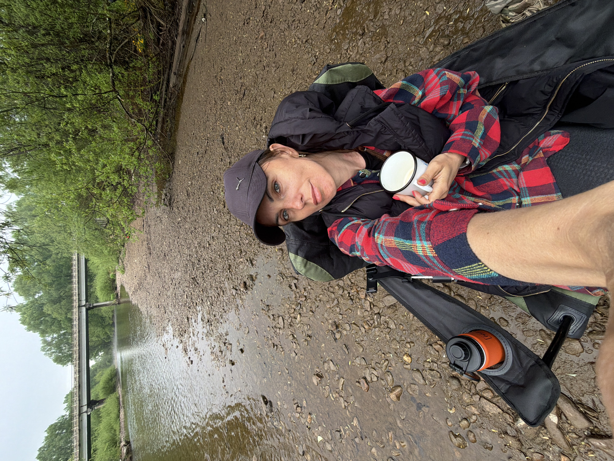 The image shows a woman sitting on a collapsible chair in a shallow, rocky stream. She wears a black baseball cap, a black vest, and a red and blue plaid shirt. She is holding a white enamel mug in her hand. Next to the chair is an orange cylindrical container in a black carrier. The water is clear and flowing. In the background is a pipeline bridge crossing the stream. The vegetation along the banks is green. The weather appears overcast. Given that location is required to be Trek, Russia, this scene could be the banks of the Snegiryovka River, where a similar pipeline bridge is located. The gravel is light, and round which is common in rivers in the region.