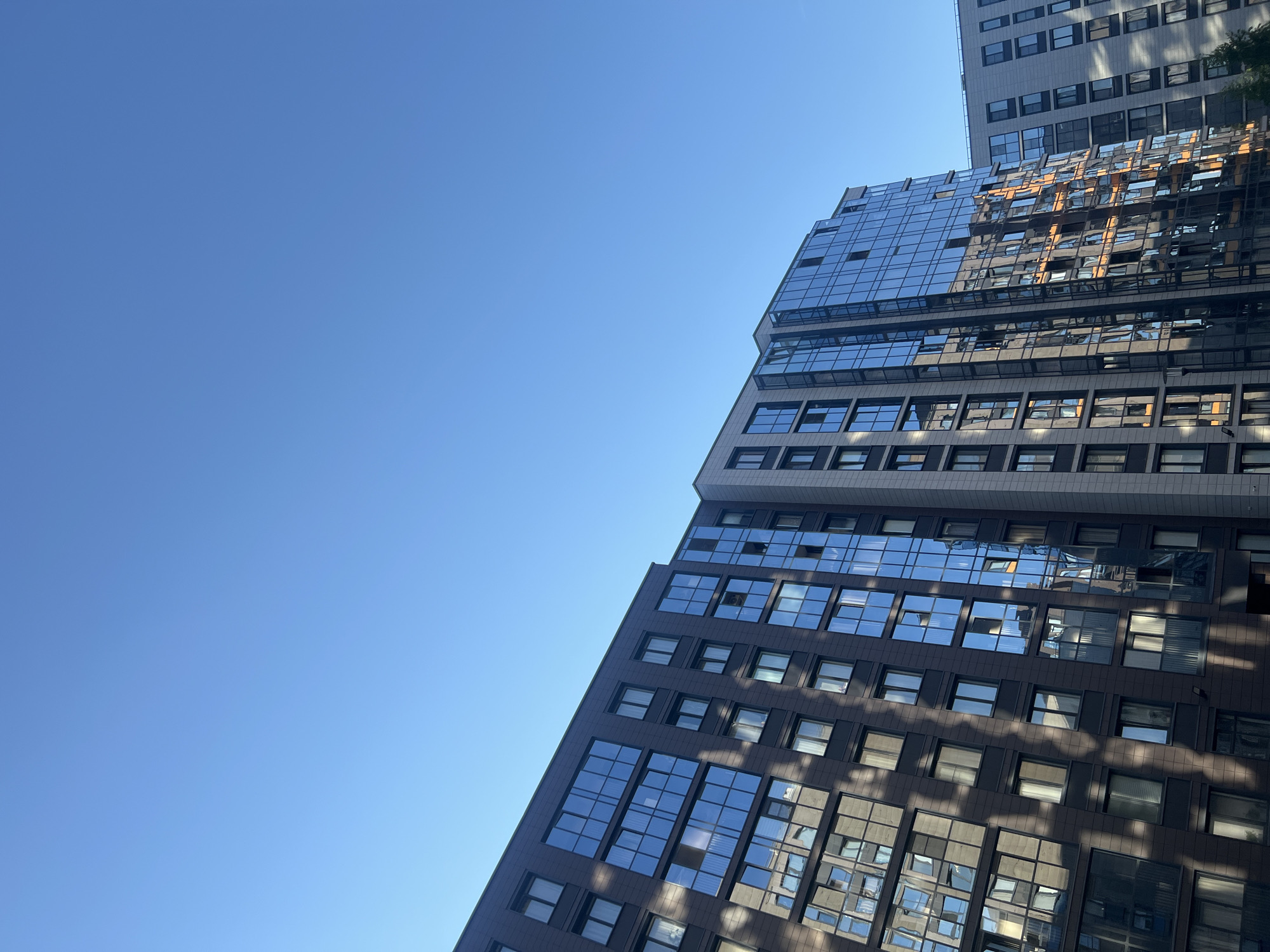 The image captures an upward-looking perspective of modern high-rise buildings against a clear, bright blue sky. The composition is divided diagonally, with the expansive, unblemished blue sky filling the upper-left portion and the architectural structures dominating the lower-right.
The buildings showcase contemporary design, featuring numerous rectangular windows and large sections of reflective glass facades. The main building in the foreground has a dark, possibly charcoal or brown, facade with rows of highly reflective blue-tinted windows. These windows reflect parts of the sky, adjacent structures, and abstract patterns of light and shadow. Behind and slightly to the right, a taller, lighter-colored building with a grid of smaller windows is partially visible. The interplay of light creates dynamic reflections and distinct shadows across the building surfaces, suggesting a sunny day with the sun at a specific angle.
The sky is a uniform, vibrant blue, completely free of clouds, indicating clear weather. No people or specific activities are depicted, giving the scene a static and architectural focus. There is no visible text on any of the buildings or in the sky. The overall impression is one of modern urban architecture, characterized by geometric lines, reflective surfaces, and the contrast between the man-made structures and the natural blue sky, typical of a bustling city environment, consistent with an unknown urban location in Russia.