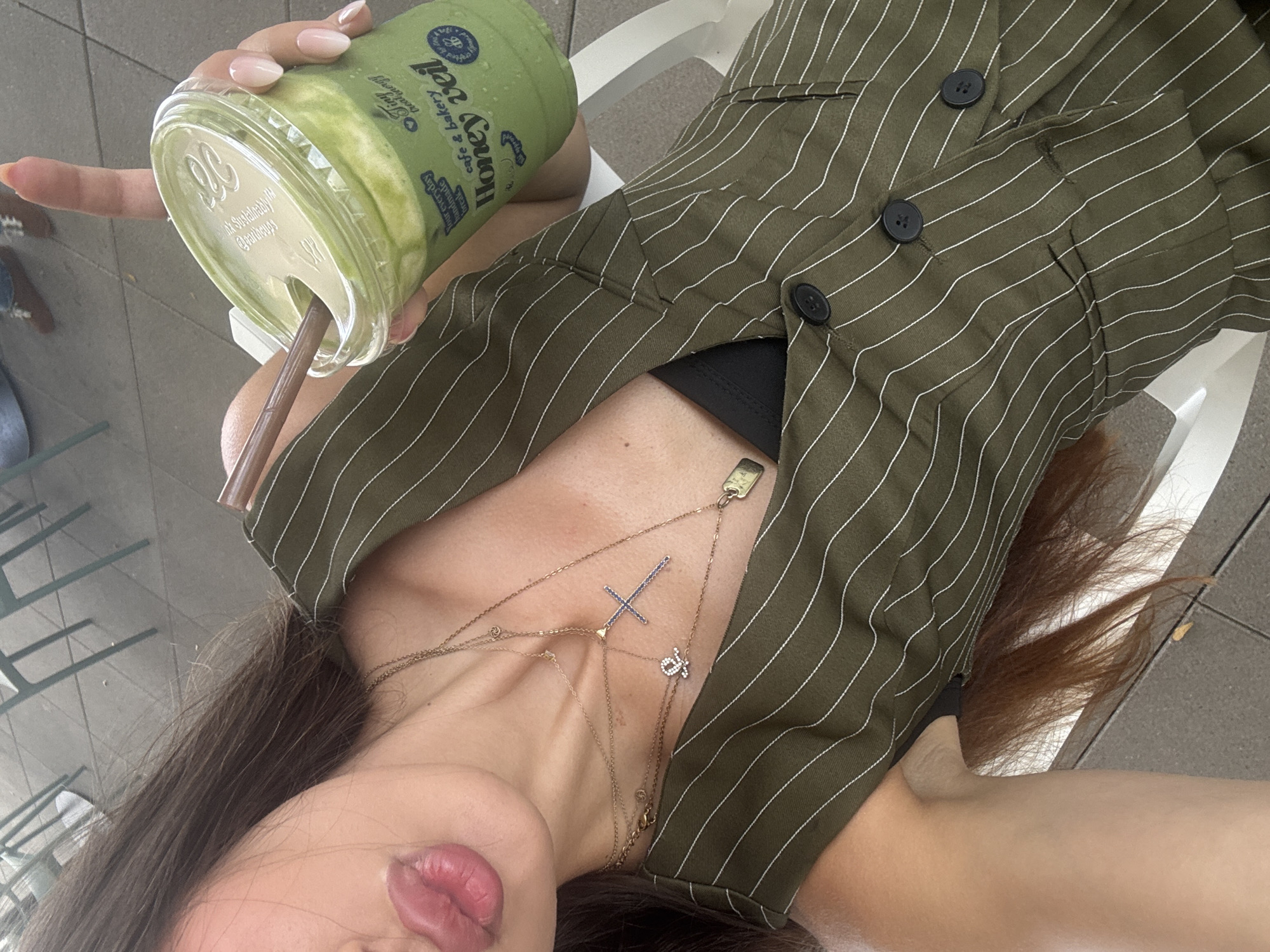 The image shows a person holding a green smoothie in a clear plastic cup. The cup has a white lid and a brown straw. The person is wearing a green and white striped jacket. There are several necklaces visible on the person's chest, including one with a cross pendant. The background suggests an outdoor setting, possibly a cafe or restaurant, with some seating visible. The lighting indicates it could be daytime. Text visible on the cup reads "Honey Veil cafe & bakery" and "Your everyday meals".