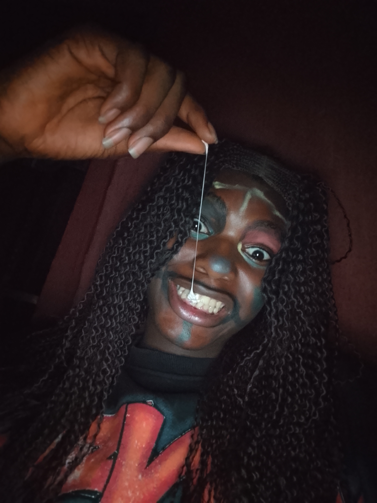 A medium-shot portrait captures a person with dark skin, long, dark, tightly curled or braided hair, and an expressive, painted face. The person is looking directly at the viewer with wide, open eyes and a broad, toothy smile. Their face is adorned with multi-colored face paint, featuring dark green/blue around the eyes and cheeks, light green/yellow lines on the forehead and nose, and hints of pink/red around the eyes.
A hand, presumably belonging to the subject, is raised into the frame from the top left, holding a thin, white, viscous string-like substance. This string stretches from between two fingers of the hand down towards the person's mouth area, creating a playful or exaggerated effect. The fingernails on the visible hand appear natural and unpolished.
The person is wearing a dark, possibly black, long-sleeved top or sweater. A portion of a red and black graphic design is visible on the chest of the garment, which appears to be the stylized logo of the band "KISS." The background is dark and indistinct, suggesting an indoor setting with dim lighting, possibly a reddish-brown wall or fabric behind the subject. The overall mood is theatrical and lively, possibly for a costume, performance, or event.