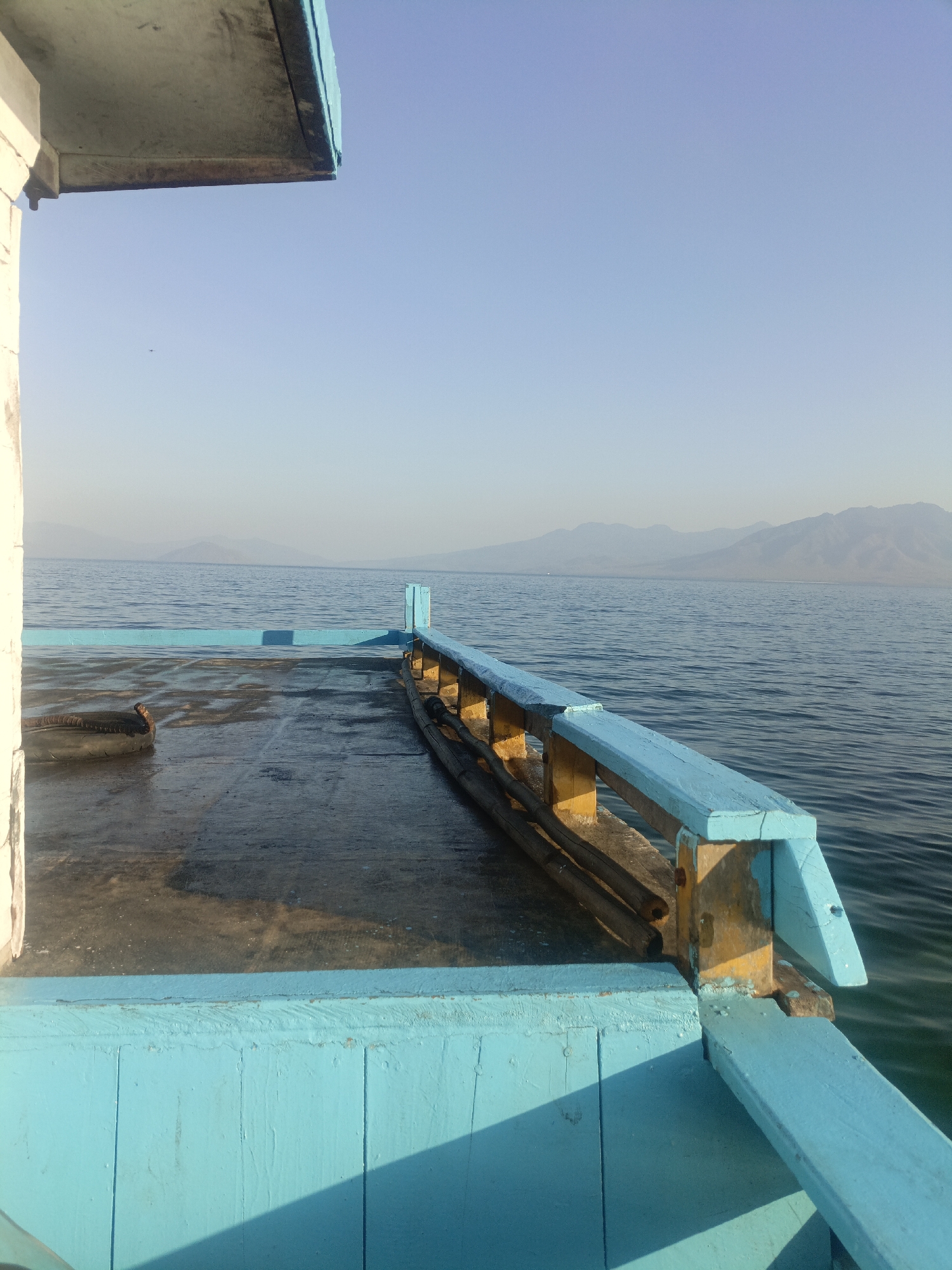 A partially visible boat deck with turquoise railings is in the foreground, with a calm body of water stretching to the horizon in the background. Mountains are visible in the distance. A dark, cylindrical object, possibly a tire, rests on the wet deck. The sky is clear and pale blue. The image appears to be taken from the perspective of someone standing on the boat. The scene is set in Ritaebang, Indonesia.