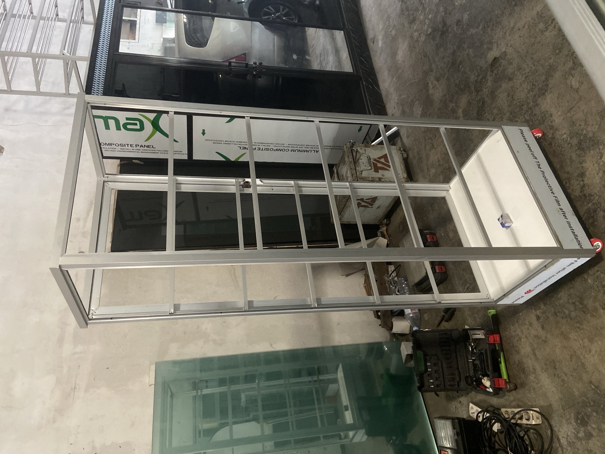 The image depicts an unfinished aluminum display case frame situated indoors, possibly in a workshop or storage area. The frame is tall and rectangular, with multiple shelves integrated into its structure. The base of the frame is white, and it is equipped with red casters for mobility. A roll of protective film with text is visible on the lower part of the frame.
In the foreground, on the left side, a black toolbox is open, revealing various tools. Electrical cords and a power strip are also present on the floor. Behind the toolbox, a large sheet of green-tinted glass is leaning against a wall, with its frame visible.
The background features a dark, enclosed structure with glass panels, possibly another display case or a storage unit. A white vehicle is partially visible through the glass of this structure. The walls appear to be concrete or plaster and show signs of wear. The overall environment suggests an active workspace where display units are being assembled or stored. The specific location of Plosokandang, Indonesia, is not explicitly indicated within the image itself.