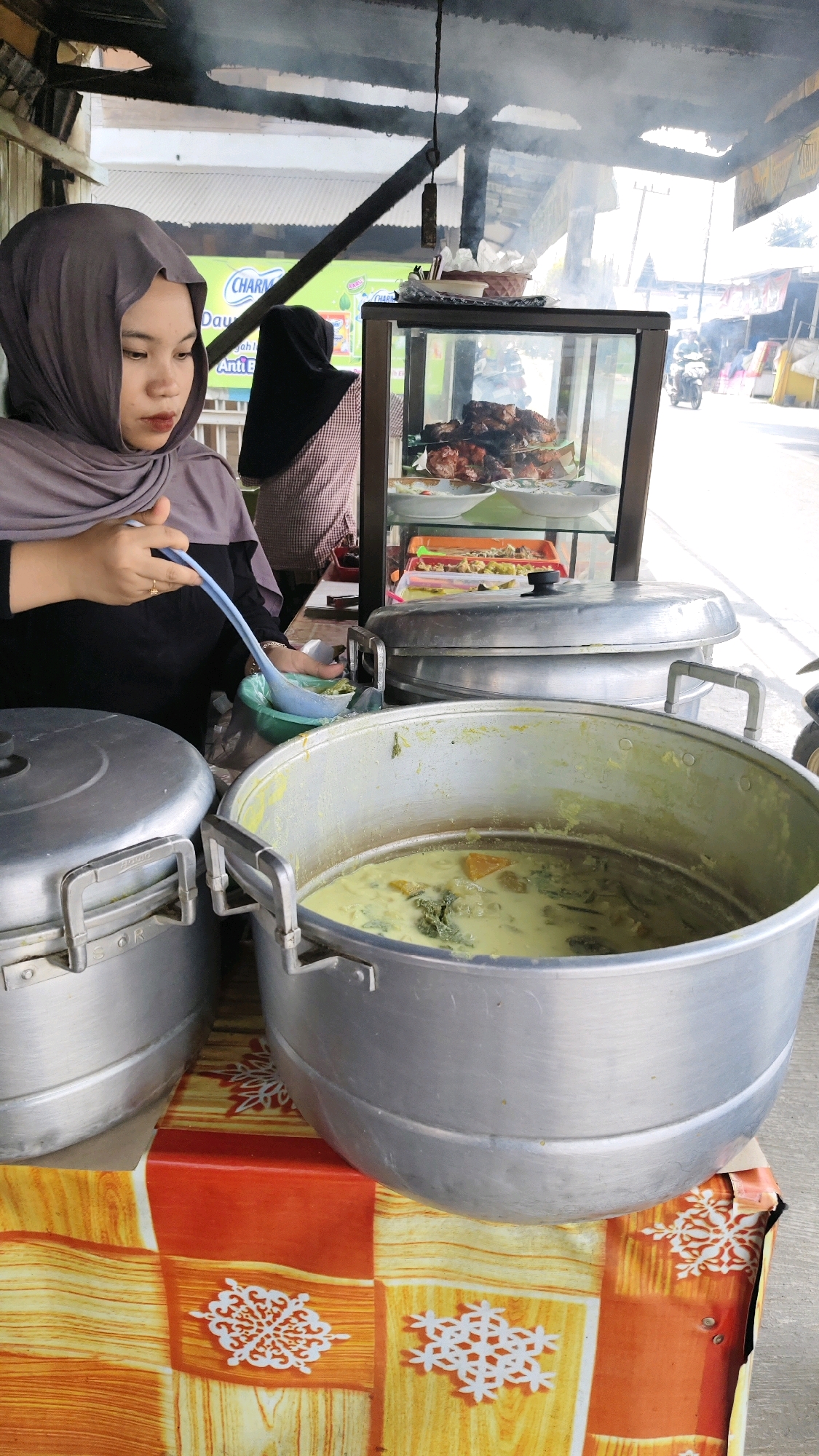 A young woman wearing a grey hijab and a black top is standing behind a food stall in Sungai Bakung, Indonesia. She is holding a ladle and is about to serve a dish from a large metal pot filled with a yellow, creamy soup containing vegetables and pieces of pumpkin. In front of the pot is another large metal pot with its lid on. Behind the woman, in a glass display case, are trays of cooked food, including grilled meat and what appears to be a curry. The stall is covered by a wooden roof, and smoke is rising, suggesting that food is being cooked. The background shows a street scene with buildings and people on motorcycles, indicating an outdoor market or roadside eatery. The table in the foreground is covered with a colorful cloth.