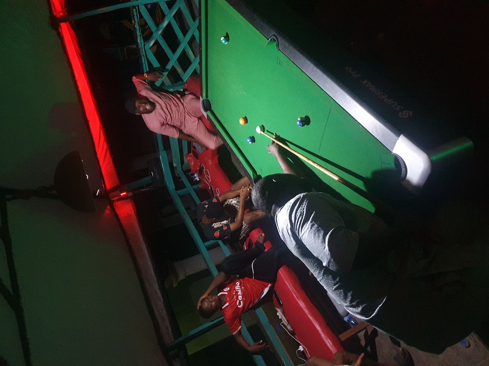 On an outdoor balcony in Lokoja, Nigeria, several individuals are gathered around a billiards table. The table is a "SUPERMAX PRO" model and has several billiards balls on the green felt surface. One person is taking a shot with a cue stick, while the other individuals appear to be observing or waiting their turn. The balcony has green wooden railing and is illuminated by a red light. The overall lighting is dim.