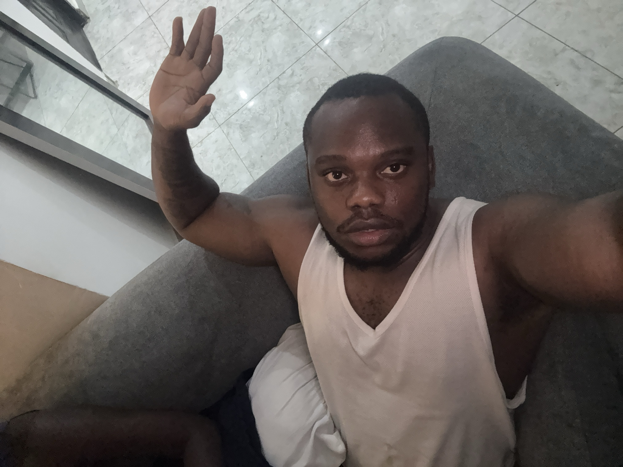 A person with dark skin is seated indoors, wearing a white tank top, and raising their left hand in a waving gesture. The person has short, dark hair and a beard. The person is seated on a gray upholstered sofa. The floor is covered in light gray tile. A window with gray framing is visible in the upper left corner of the image. There is insufficient information to determine the specific location of the subject within Iddo Tudunwada, Nigeria.
