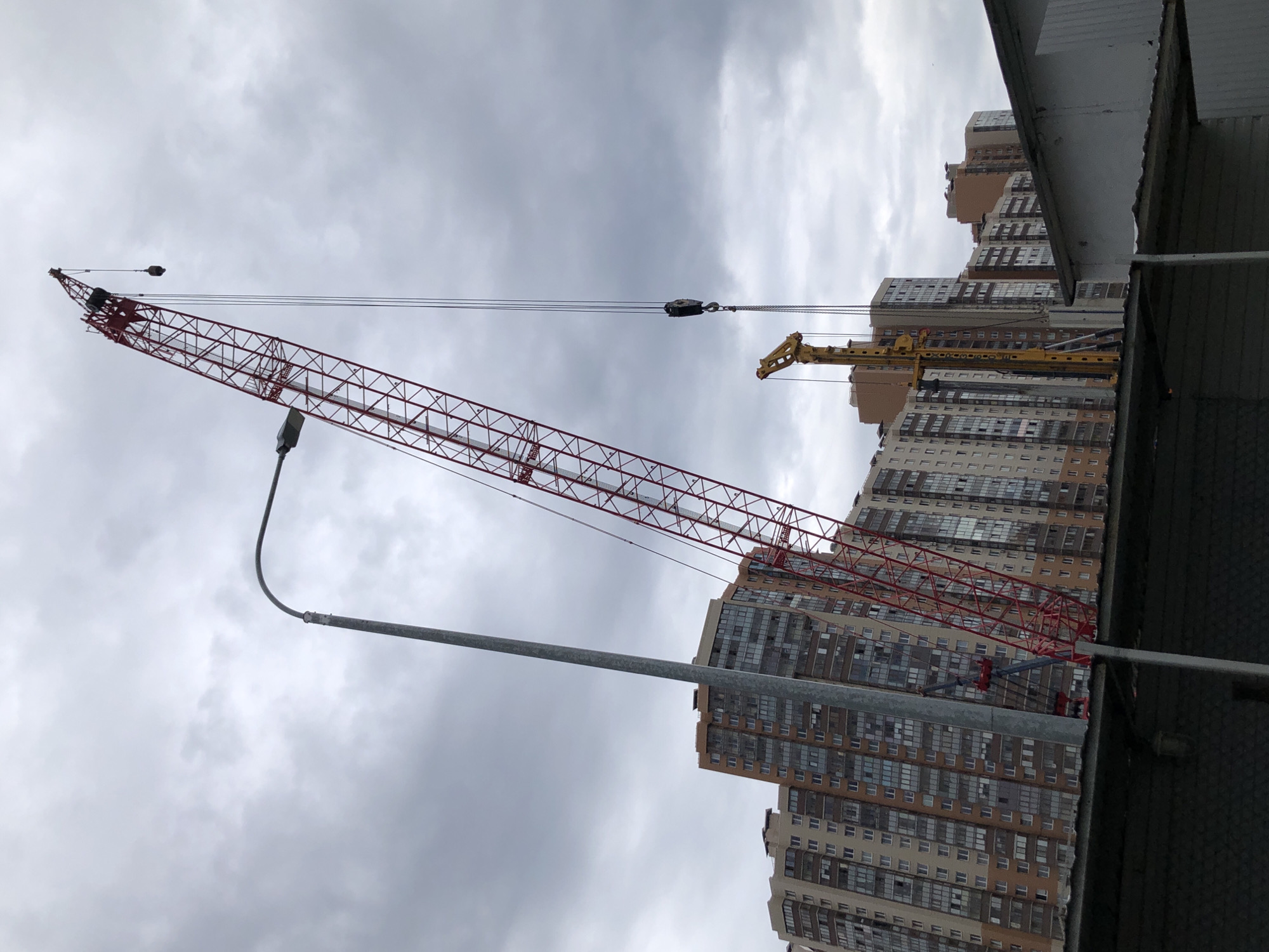 A construction scene unfolds in Saint Petersburg, Russia, under a cloudy sky. A massive red lattice boom crane dominates the frame, its long boom extending upwards and to the left, with cables and a hook suspended from its peak. To the left of the crane, a tall, slender lamppost with a curved arm arcs towards the sky. In the background, a series of tall, modern residential buildings, characterized by their numerous windows and light-colored facades with reddish-brown accents, rise in neat rows. A yellow piling rig, another piece of construction equipment, stands to the right of the lamppost and in front of the buildings. In the foreground, the dark, corrugated roof of a low-lying structure, possibly a temporary building or shed, partially obscures the lower portion of the scene, creating a sense of depth and perspective. No people are visible in the frame.