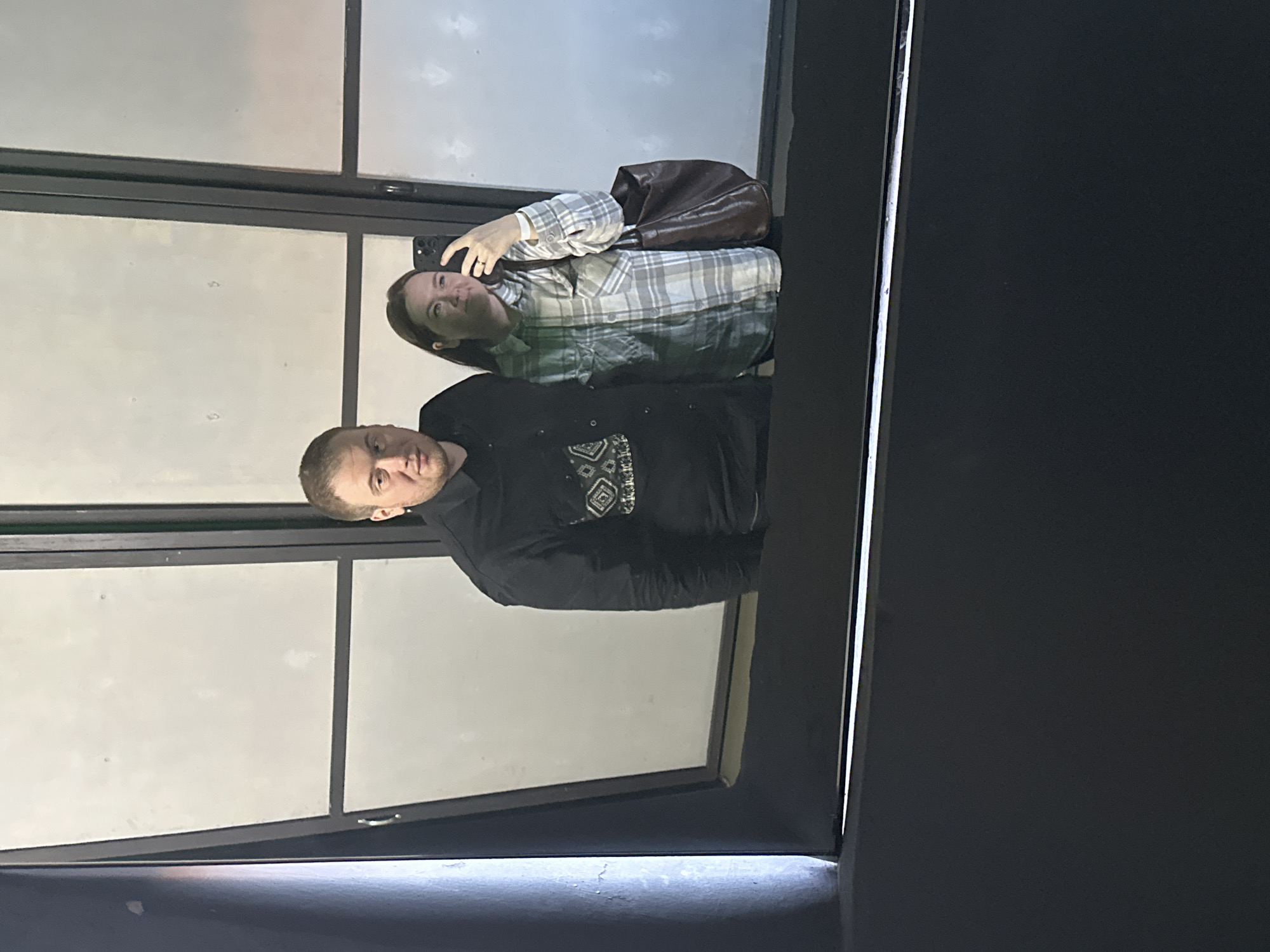 A man and a woman are reflected in a window. The woman is holding a cell phone and appears to be taking a picture of the reflection. The man is standing next to her. The location is not discernible from the image.