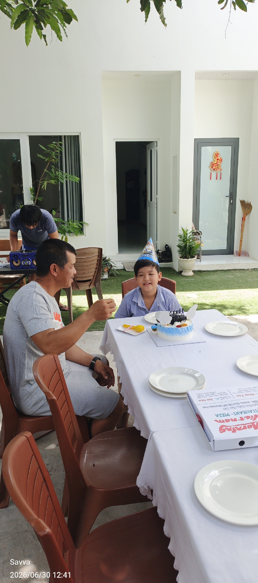 Two males, an adult and a child, are seated at a white-clothed table outdoors in a courtyard. The adult, on the left, wears a grey t-shirt and grey shorts and is holding up a small object towards the child. The child, on the right, wears a light blue collared shirt and a blue party hat. A small cake with blue and white frosting and black decorations sits on the table in front of the child. There are white plates on the table and a pizza box is visible. In the background, there is a white building with open doorways and a grey door with red decorations. Green grass covers the ground. The scene is likely a birthday celebration.