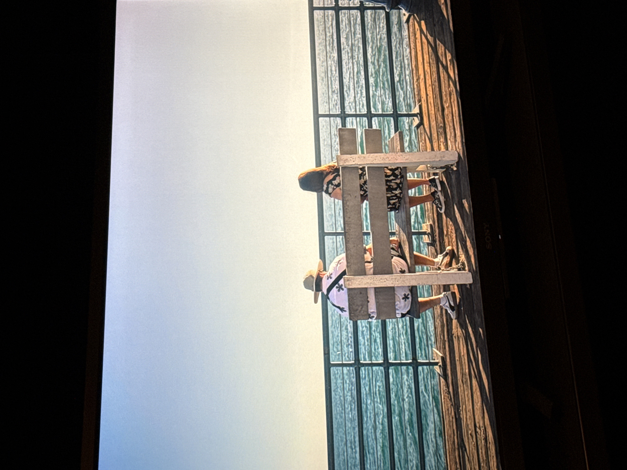 Two individuals are seated on a bench at the edge of a pier. A body of water is seen through a metal fence. The pier's structure appears to be constructed of wood. The location is likely coastal, but not specifically identifiable as being in London.