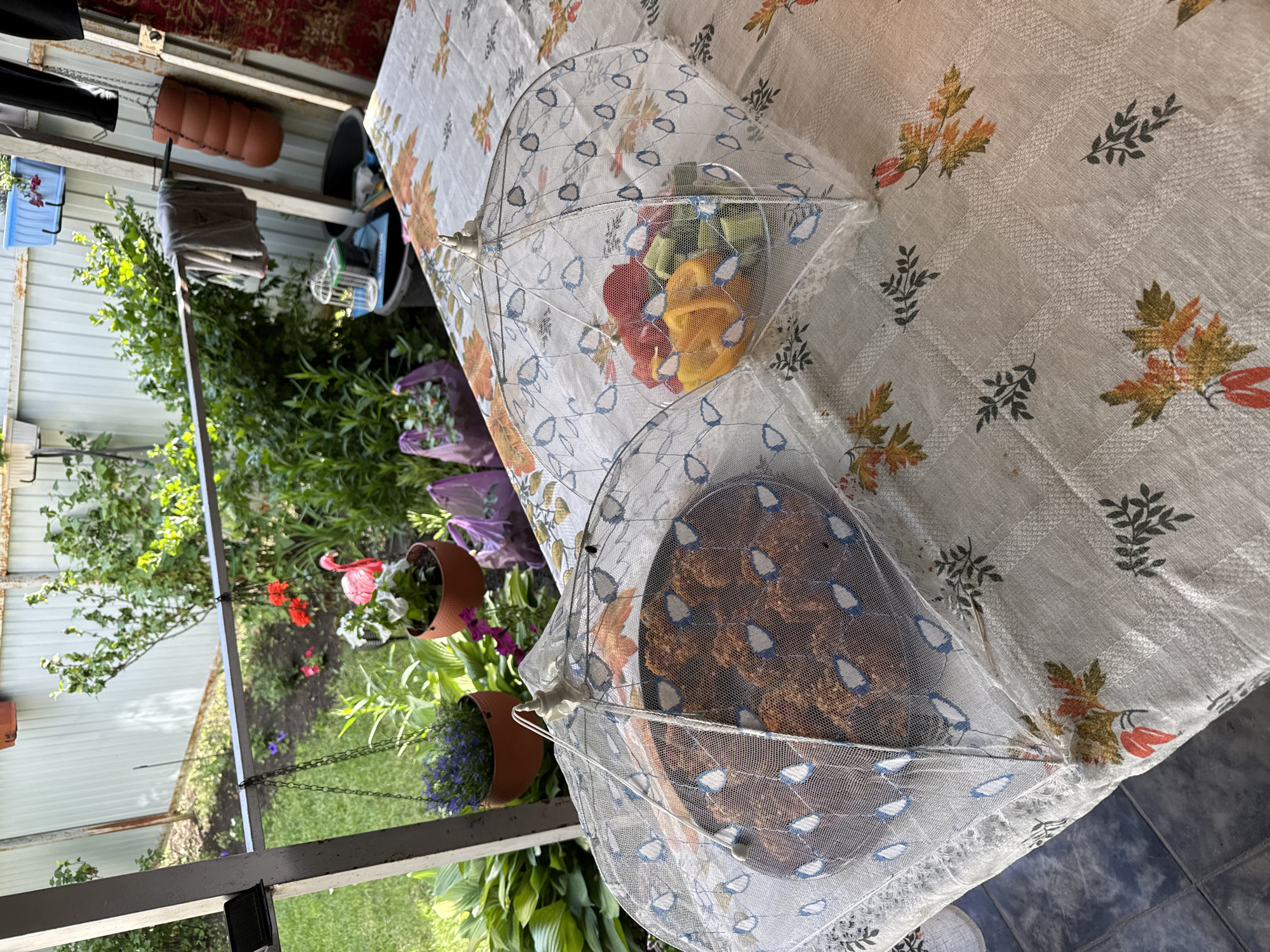 The image shows an outdoor table covered with a floral-patterned tablecloth. On the table are two food covers, one containing a platter of fried food items, and the other containing chopped vegetables such as bell peppers and cucumbers. In the background, a garden area with greenery and flowers is visible, along with a fence. The setting appears to be a backyard or patio area, likely in a rural or suburban location given the presence of the garden and fence. There are no people visible in the image. The overall impression is one of an outdoor meal preparation or serving scene.