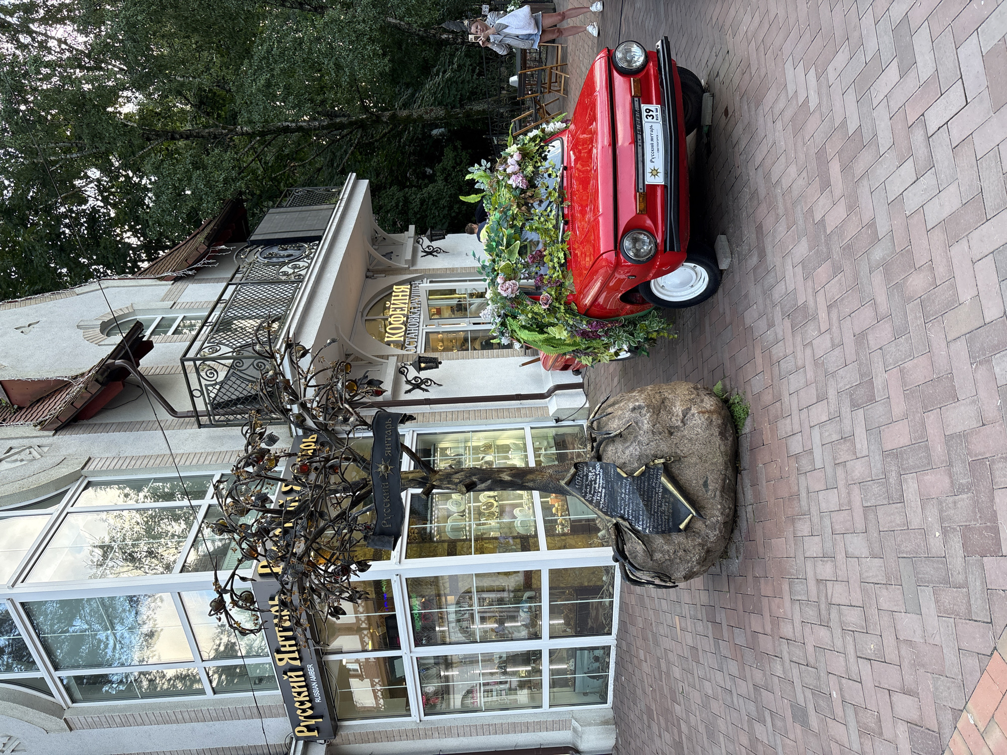 In the city of Svetlogorsk, Russia, a red vintage car, decorated with an abundance of flowers and greenery, is parked on a brick-paved street in front of a building with large windows and ornate balconies. A woman stands to the right of the car, holding a smartphone and taking a picture. To the left of the car, a sculpture of a tree with metal branches and amber-like ornaments is mounted on a large boulder with an inscribed plaque. The building behind the car has signage indicating a jewelry store named "Russian Amber" and a cafe. The scene is set against a backdrop of lush green trees, suggesting an outdoor public space.