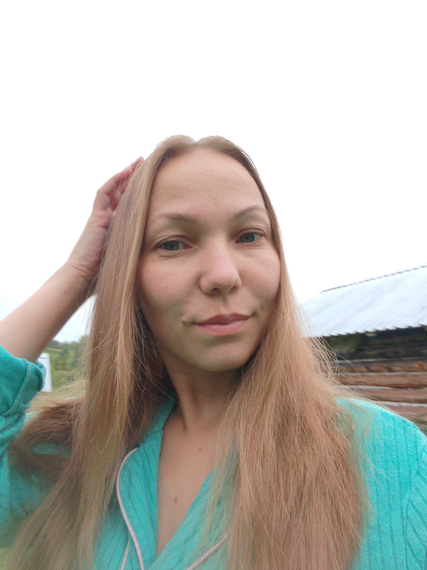 The image is a close-up, front-facing selfie of a woman with long, light brown hair, taken outdoors. She is wearing a teal-colored garment with pink trim. Her left hand is raised and touching her hair. In the background, there is a wooden structure with a white roof, suggesting a rural or rustic setting. The sky is overcast and bright white. Based on the provided context, the location is identified as Чухлэм, Russia.