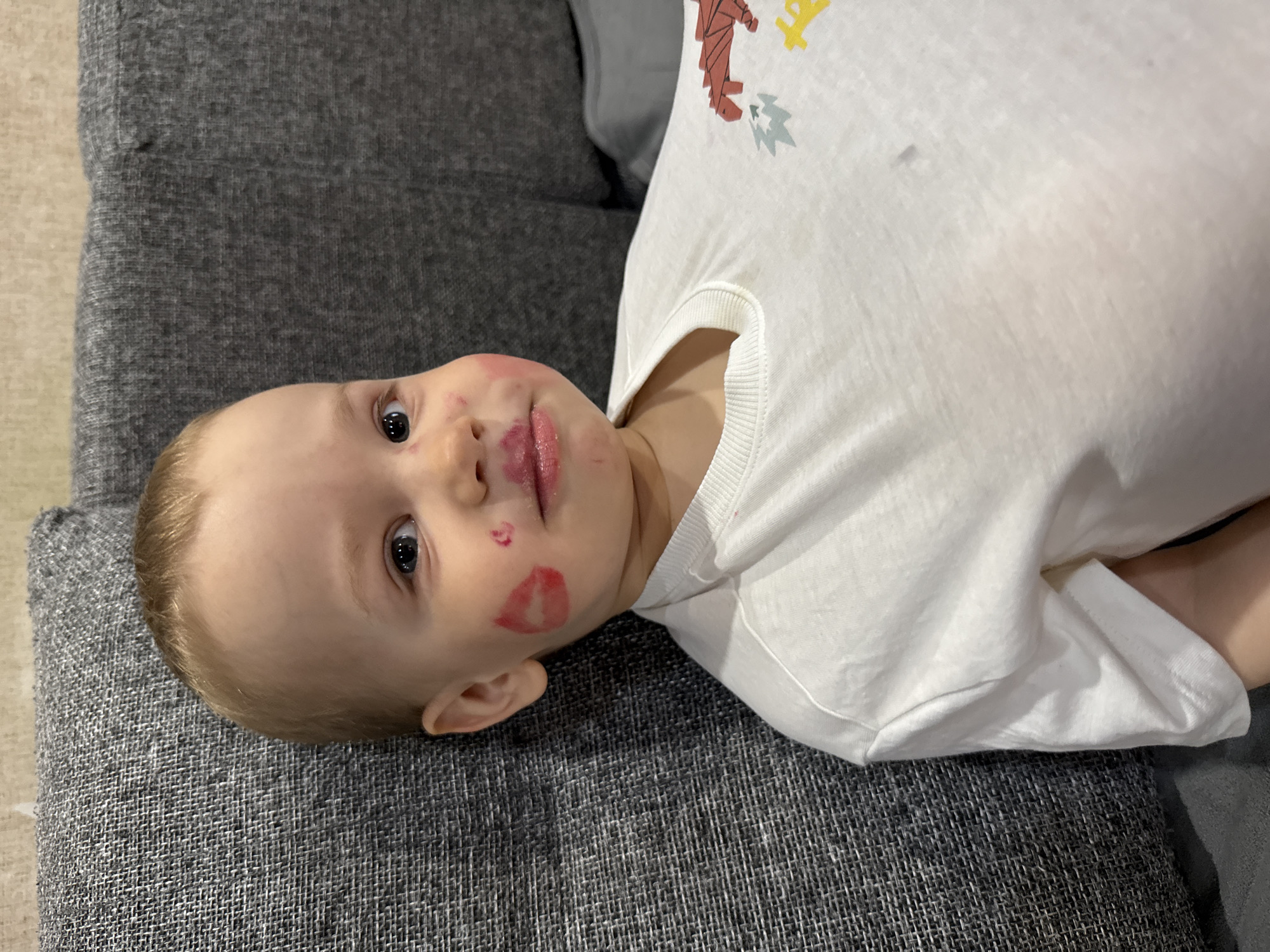 A young child with blond hair is lying on a grey sofa. The child is wearing a white shirt with a design on the upper right portion. Red lipstick marks are visible on the child's lips and cheek. The location is possibly Rusanovo, Russia, but there are no elements in the image to confirm this.