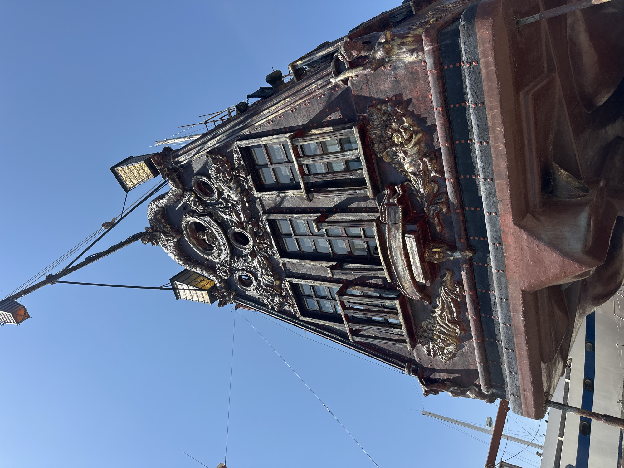 The image shows the stern of a replica of a historical sailing ship, possibly a galleon, in Bar, Montenegro. The ship features ornate, dark red and gold decorations, including carved figures, elaborate scrollwork, and multiple windows. The vessel is partially elevated on supports, with other modern ships visible in the background, suggesting a shipyard or harbor setting. The sky is clear and blue.