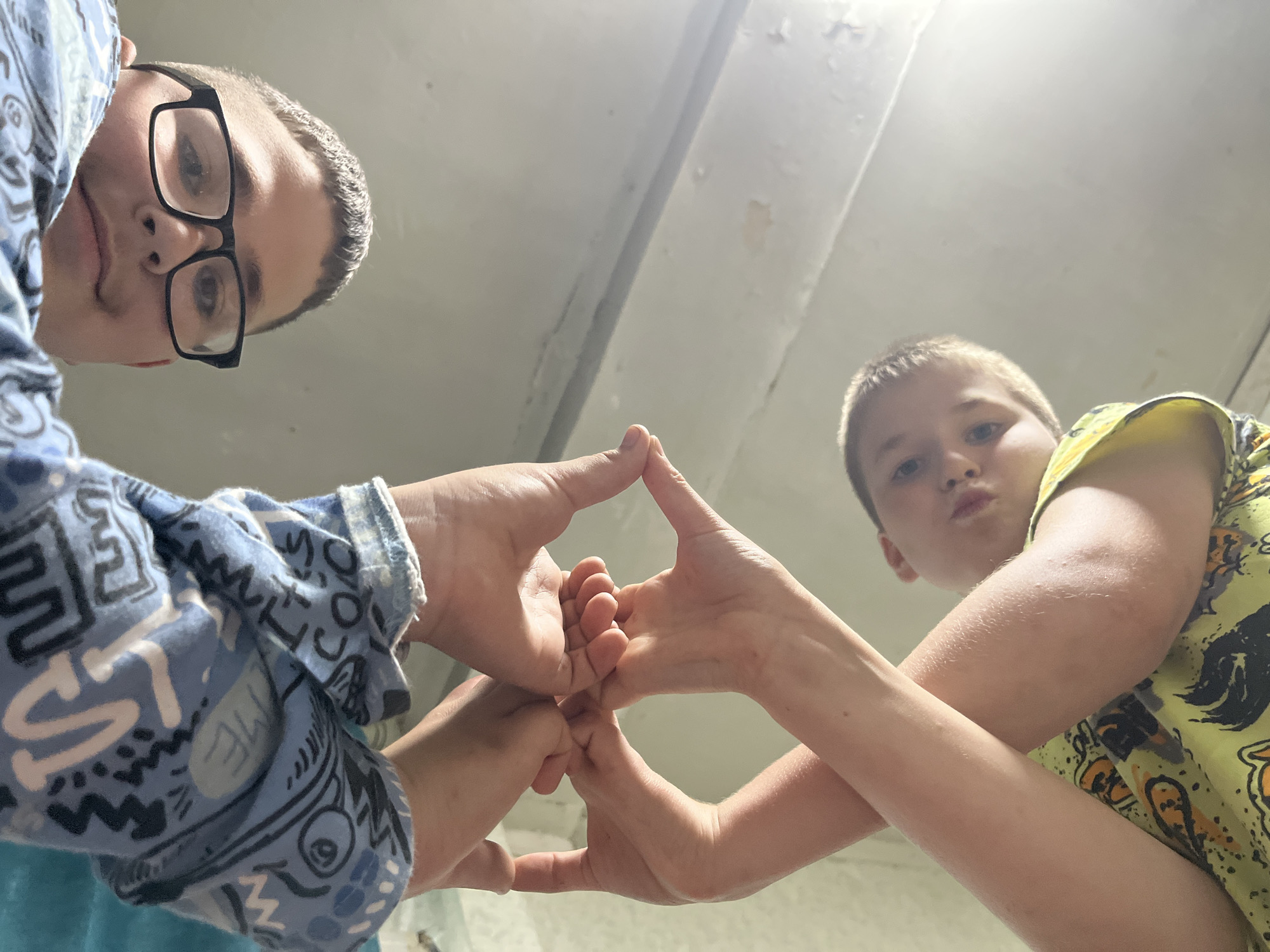 The image shows two young boys looking up at the camera from a low angle. The boy on the left is wearing glasses and a blue shirt with a repeating graphic pattern. The boy on the right is wearing a yellow t-shirt with an abstract design. Both boys are holding their hands up, forming a diamond shape with their fingers. Their faces are partially obscured by the angle of the shot. The background appears to be an indoor space with a ceiling that shows signs of wear, including peeling paint. The lighting suggests it may be daytime, possibly indoors with artificial lighting. The overall mood is playful and cooperative, as indicated by the hand gesture and the shared activity. No specific time of day or weather conditions are discernible. The location context provided is Фоки, Russia, but there are no explicit visual cues in the image to confirm this. There is no visible text in the image.