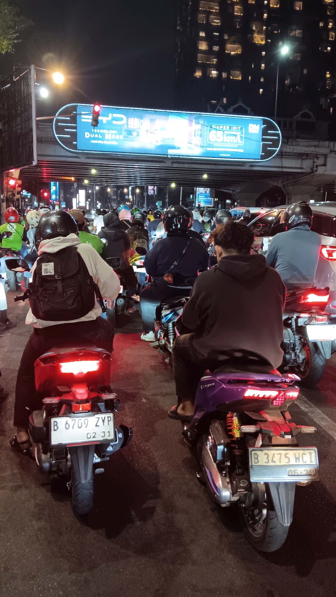 The image depicts a traffic jam at night on a city street in South Jakarta, Indonesia. A large, illuminated billboard with the text "DYP TEKNOLOGI DUAL MODE" and "SUPER IRIT 65 KM/L" is visible overhead, spanning across the road. Numerous motorcycles are stopped in traffic, with their riders wearing helmets. The foreground shows the rear view of two motorcycles. The motorcycle on the left has a license plate that reads "B 6709 ZYP 02-31". The motorcycle on the right has a license plate that reads "B 3475 WCI 06-29". The road surface is dark and appears wet in some areas, reflecting the lights. In the background, tall buildings with illuminated windows are visible, suggesting an urban environment. The overall scene is characteristic of a busy city street at night, with traffic congestion and illuminated advertising.