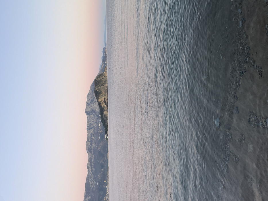 The image presents a wide coastal landscape from Budva, Montenegro, captured during twilight hours (dawn or dusk). The immediate foreground shows transparent seawater over a sandy seabed intermixed with dark, irregular rocks and pebbles. The water surface extends across the midground with visible ripples, reflecting the ambient light. In the background, a range of dark, undulating mountains occupies the horizon. A prominent, rocky landmass, identifiable as Sveti Nikola Island, is situated in the mid-right, extending into the water. Along the base of the mountains, scattered buildings and faint artificial lights indicate distant coastal settlements, with a denser cluster visible to the far left. The sky transitions from a light blue at the top to soft pink and peach tones closer to the horizon. A spherical, yellowish-orange light source is visible low on the horizon to the far left, behind the distant landmass. No human figures or vessels are present within the frame.