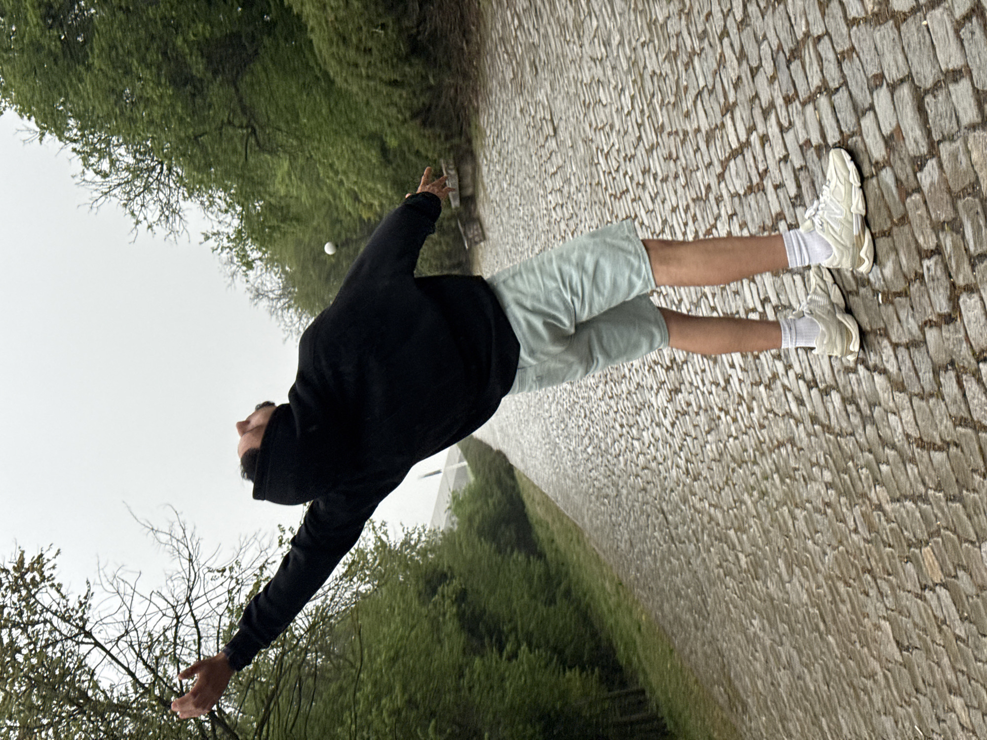 A man stands on a cobblestone path, throwing a small, white ball into the air. He is wearing a black hooded sweatshirt, light green shorts, white socks, and white sneakers. His arms are outstretched, with his right arm and hand positioned above his head to throw, while his left arm is extending outward. The man is looking up.
The background consists of lush green trees and foliage on either side of the path. The sky is overcast and grey.
The image was taken in Shumen, Bulgaria.