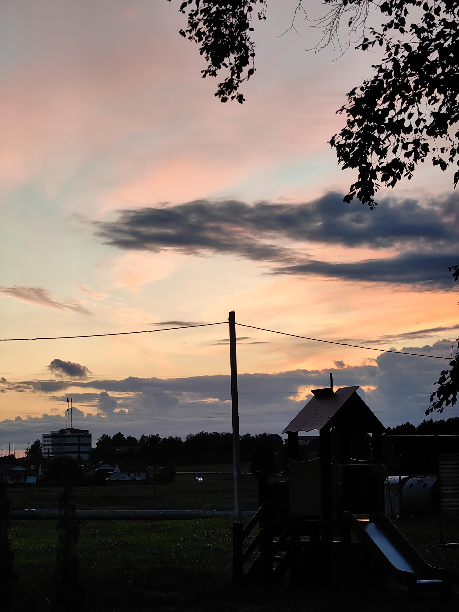 The image depicts a silhouetted playground structure in the foreground, featuring a slide and climbing elements. Behind the playground, a field stretches out, with a road or path visible. In the middle ground, several houses and trees are present. To the left, a tall, multi-story building with antennas on top stands out, suggesting an urban or suburban fringe. The background is dominated by a dramatic sunset sky, with bands of pink, orange, and gray clouds. Tree branches, also silhouetted, frame the top of the image. The scene appears to be set in a location with open space, likely on the outskirts of a town, potentially Myadzyel, Belarus, given the context provided. No people or animals are visible in the frame.
