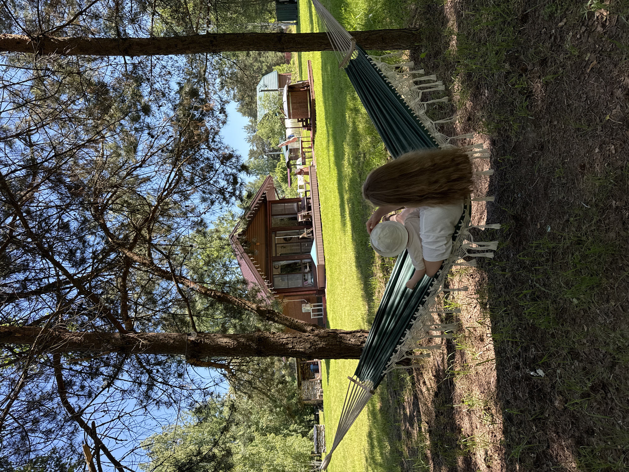 This detailed description outlines a sunny outdoor leisure scene located in Копнино, Russia.
**Scene Description:**
The image depicts a person relaxing in a dark green and white striped hammock, suspended between two prominent pine trees, observed from a low-angle rear perspective. The individual has long, light brown hair and is wearing a light-colored top and a light-colored bucket hat, with their right hand seemingly touching the hat. The hammock features white decorative tassels along its edges and is secured by rope to the tree trunks.
The immediate foreground under the hammock is shaded, showing bare earth with sparse patches of grass and fallen debris. Beyond this, a wide, brightly sunlit expanse of well-maintained green lawn dominates the mid-ground.
In the background, several wooden buildings are visible under a clear, bright blue sky. To the left, a rustic, log-style structure, possibly a wood shed, is partially obscured by trees. Centrally located is a two-story wooden house with dark brown siding, a reddish-brown gabled roof, and large windows or glass doors. A white exterior unit (likely air conditioning) is mounted on its side, and white string lights are draped along its eaves. A patio with outdoor chairs and a small table is visible in front of this house.
To the right of the central house, an outdoor dining area with a table and chairs is set up under a light-colored canopy. Adjacent to this, a wooden hot tub or outdoor bath structure is positioned on a wooden deck, with a small flag (partially obscured) flying nearby. Further in the background, another wooden house with a green gabled roof is visible, along with two dark green rectangular utility containers on the far right. The overall environment suggests a recreational or holiday property, characterized by natural elements and rural-style accommodations bathed in bright daylight, casting distinct shadows.
