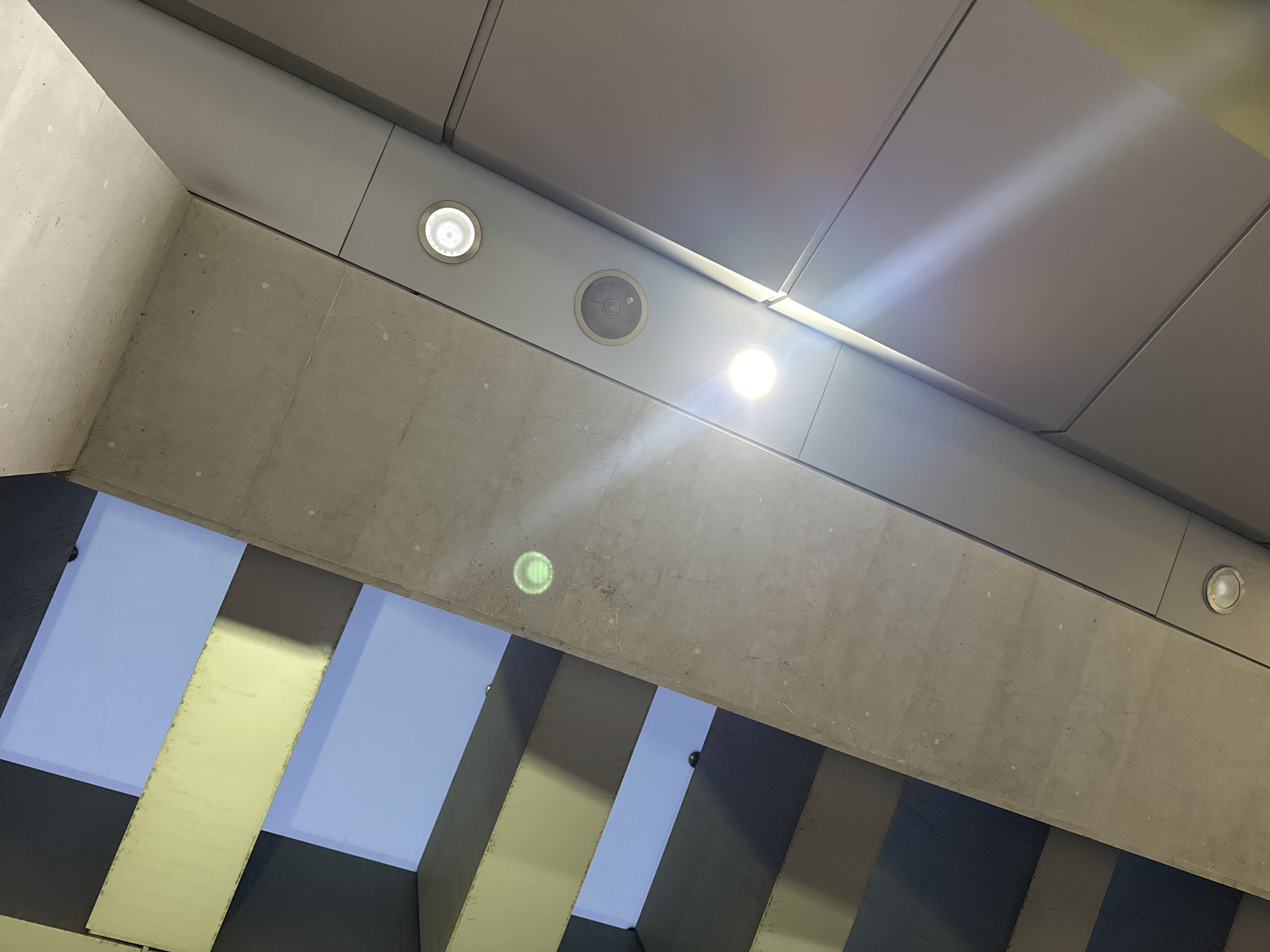 The image captures an upward-looking view of a modern architectural space, possibly an underpass or covered walkway, characterized by geometric forms and a blend of concrete and metal. The upper section features a gray ceiling composed of rectangular, modular panels. A prominent, unfinished concrete beam diagonally dissects the frame, forming a structural element.
Integrated into the ceiling and the side of the concrete beam are several circular recessed lights. Two of these lights are brightly illuminated, emitting strong white light and visible lens flares, while another light and a circular recessed speaker are also visible. A distinct, circular green reflection or light is projected onto the concrete beam. Below this main structure, multiple vertical or angled supporting beams, varying in shades of light and dark gray, are present. Between these supports, sections of a light blue background are visible, suggesting an open sky or an outdoor area beyond.
The scene is devoid of people, highlighting the clean, industrial, and contemporary design of the infrastructure. The interplay of artificial lights against what appears to be natural daylight implies a functional, possibly public, environment in Yerevan, Armenia. There is no visible text.