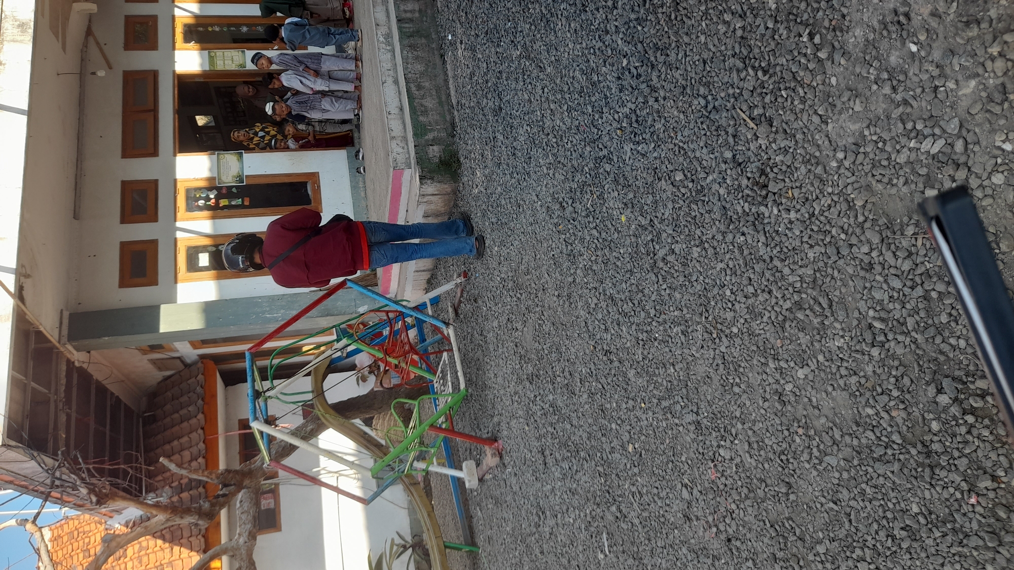 The image depicts an outdoor scene in front of a building, likely a school or community center, in Selaawi, Indonesia. In the foreground, a person wearing a maroon jacket, blue jeans, and a black helmet is standing, facing away from the camera. To the left, a colorful playground structure with swings and a slide is visible. The ground is covered in gravel. In the background, several children, dressed in light-colored Islamic attire, are gathered near the entrance of the building, some appearing to be interacting with adults. The building has a light-colored exterior with windows and an open doorway. The overall setting suggests a daytime scene with natural lighting.