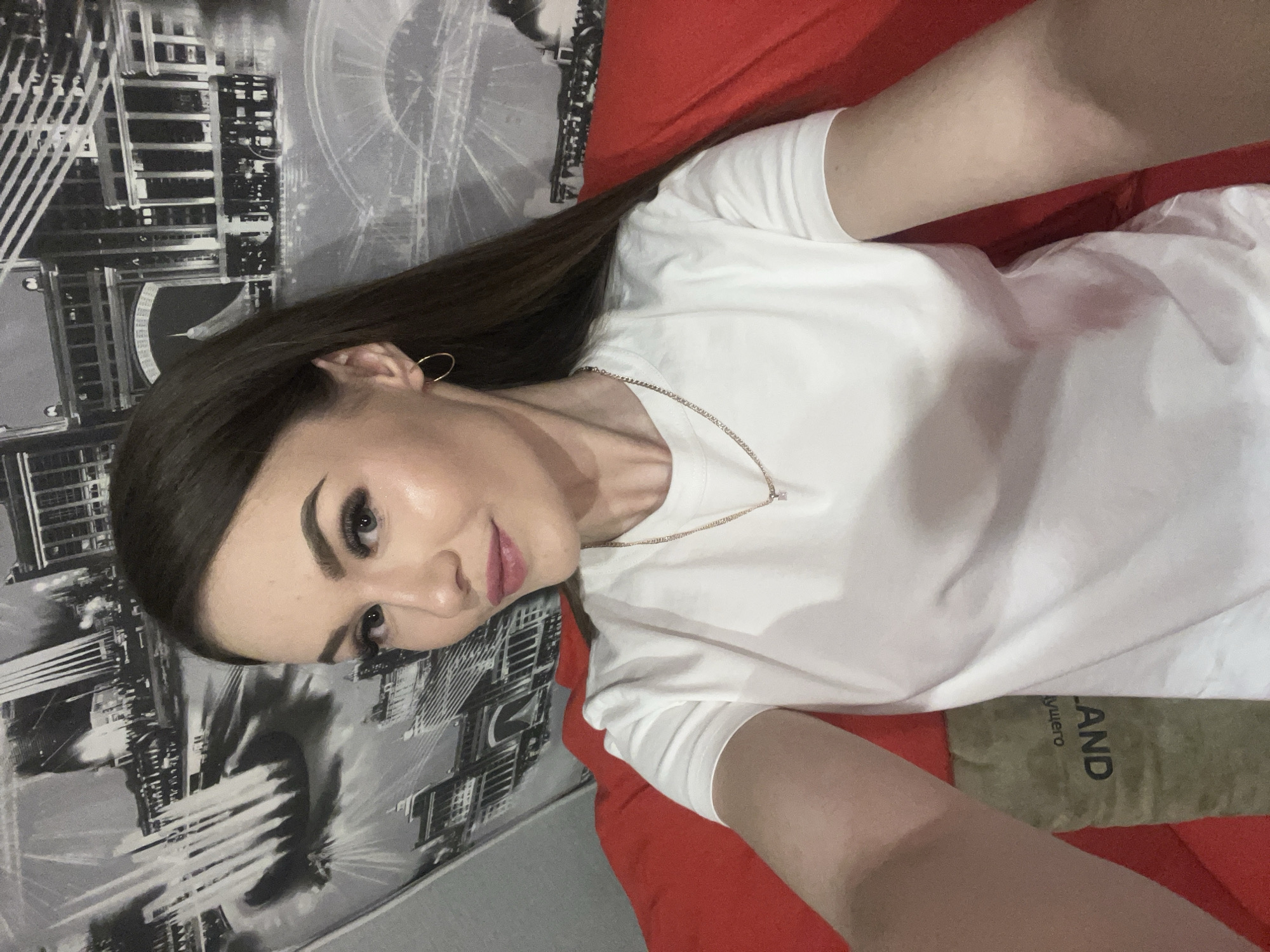 A woman with long brown hair is lying on a red surface, possibly a bed, wearing a white t-shirt and a gold chain necklace. Her head is tilted, and she is looking towards the camera with a slight smile. The background features a black and white cityscape image on the wall. A decorative pillow with text that includes the word "LAND" is partially visible on the right side of the image. The location cannot be determined.
