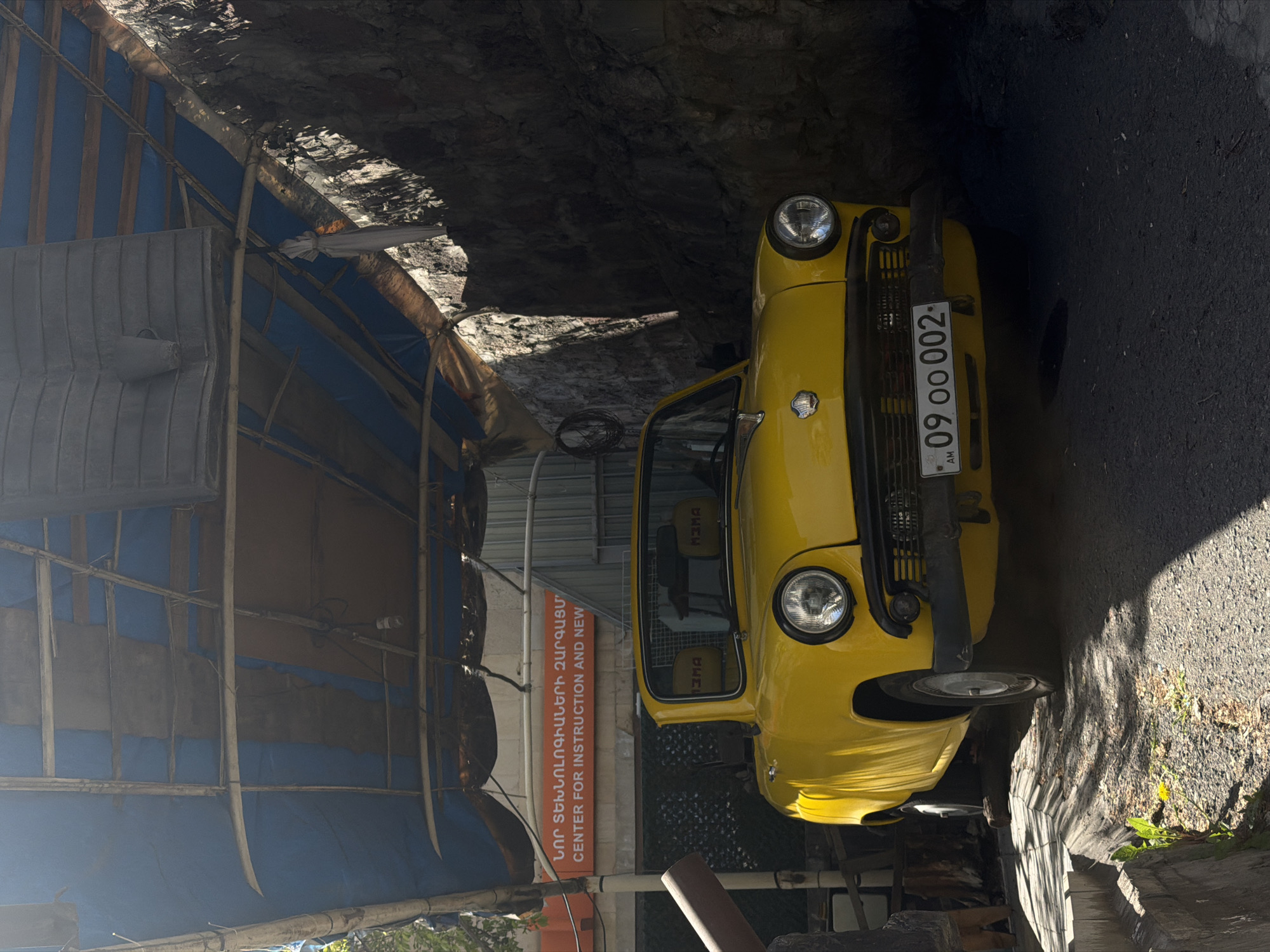 An upright view of the image reveals a bright yellow vintage car, possibly a ZAZ-965 or similar classic compact model, parked outdoors during the daytime. The car is seen from the front-right, featuring distinctive circular headlights and a chrome grille. Its Armenian license plate, "09 OO 002," is prominently displayed, showing the country code "AM." The car's interior is partially visible through the windshield, revealing patterned seats.
The setting appears to be a narrow passage or alleyway. To the right of the car, a rough, dark rocky wall or cliff face rises, showing textured surfaces with patches of sunlight highlighting details. To the left, a makeshift structure with a blue and brown tarp or fabric covering is erected, supported by a framework of poles.
An orange sign with white text is affixed to the temporary structure. The Armenian script reads "ՆՈՐ ՏԵԽՆՈԼՈԳԻԱՆԵՐԻ ԶԱՐԳԱՑՄԱՆ" (Development of New Technologies), followed by the English translation "CENTER FOR INSTRUCTION AND NEW". This suggests the full English text refers to a "Center for Instruction and New Technologies Development."
The ground is dark asphalt, with some sparse greenery, including a small dandelion, visible near the rocky wall. Strong shadows indicate a sunny day. No people or specific activities are observed; the car is simply parked in what appears to be a utilitarian or informal space within the city of Yerevan, Armenia.