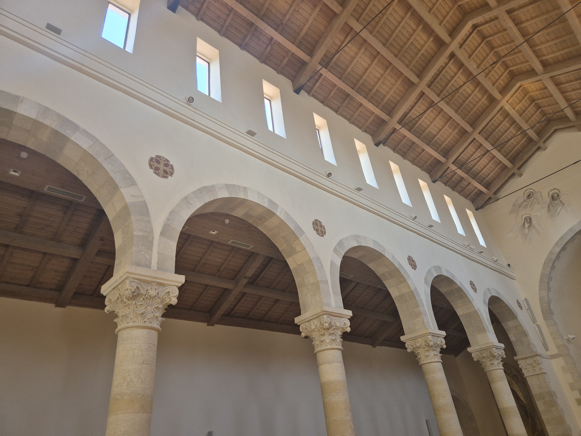 The image shows the interior of a building, likely a church or a basilica, in Abu Gosh, Israel. The architecture features a series of large stone arches supported by ornate columns. The arches are open, revealing the interior of the building with a wooden, vaulted ceiling supported by beams. Natural light streams in from tall, narrow windows positioned high on the wall above the arches. Along the wall, there are decorative circular motifs and a faded mural depicting figures, possibly angels or saints. The overall impression is one of historical and religious significance, with a focus on stone and wood construction and classical architectural elements.
