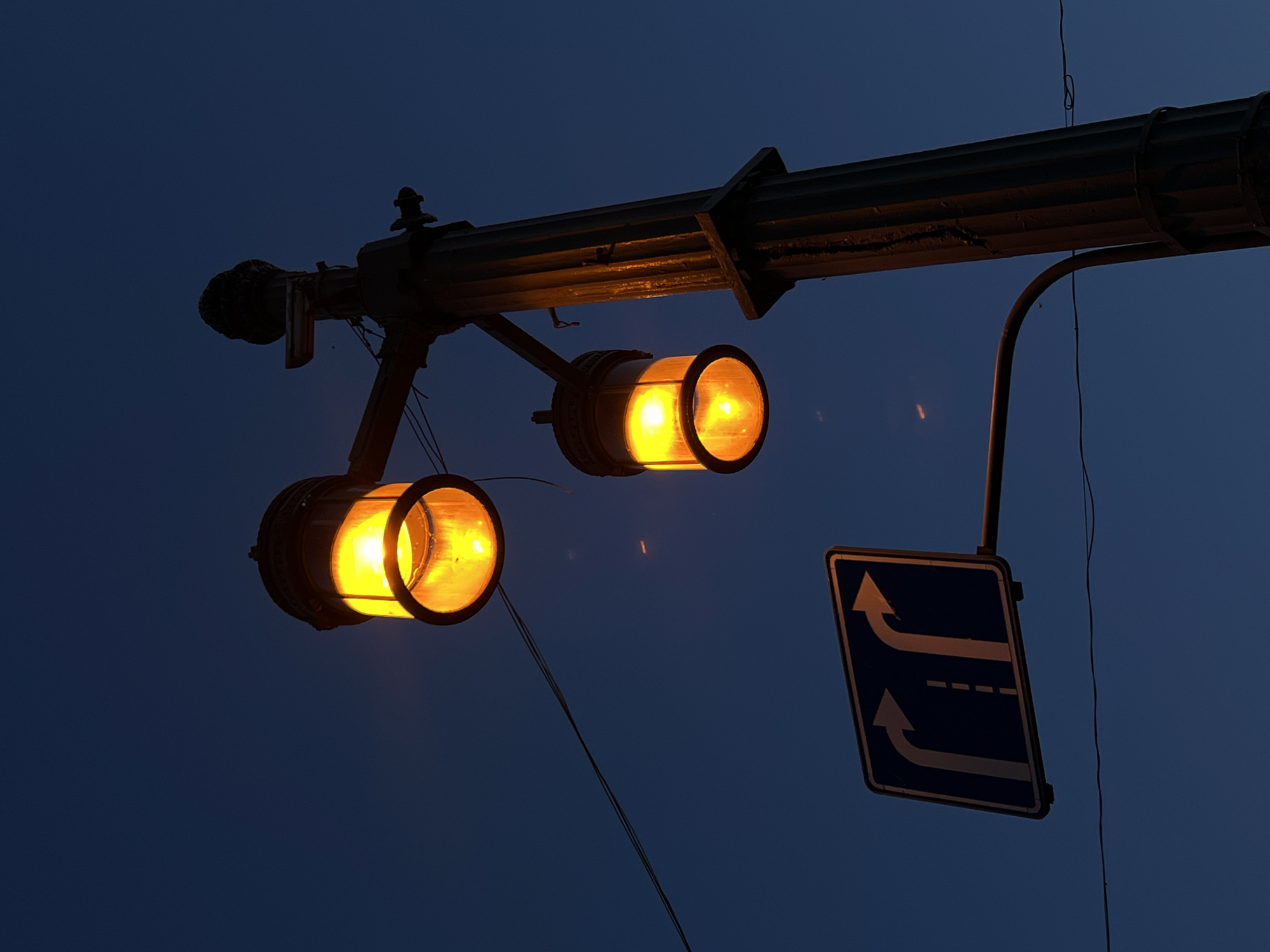 This is a low-angle, close-up shot of a street light and a directional road sign against a dark blue evening sky. The street light has two glass-encased lamps that emit a warm, yellow-orange glow. The lamps are of different sizes, with the upper one being larger and the lower one smaller. The light post itself is a dark, ornate structure with fluted detailing, casting a silhouette against the sky.
To the left of the street light, a blue rectangular road sign with white arrows is visible. The sign indicates that traffic should go straight or turn right. A thin black wire runs horizontally across the lower part of the frame. The environment appears to be an urban setting at dusk or dawn, possibly in Unknown, Russia, given the context of the original prompt. There are no people or animals visible in the image.